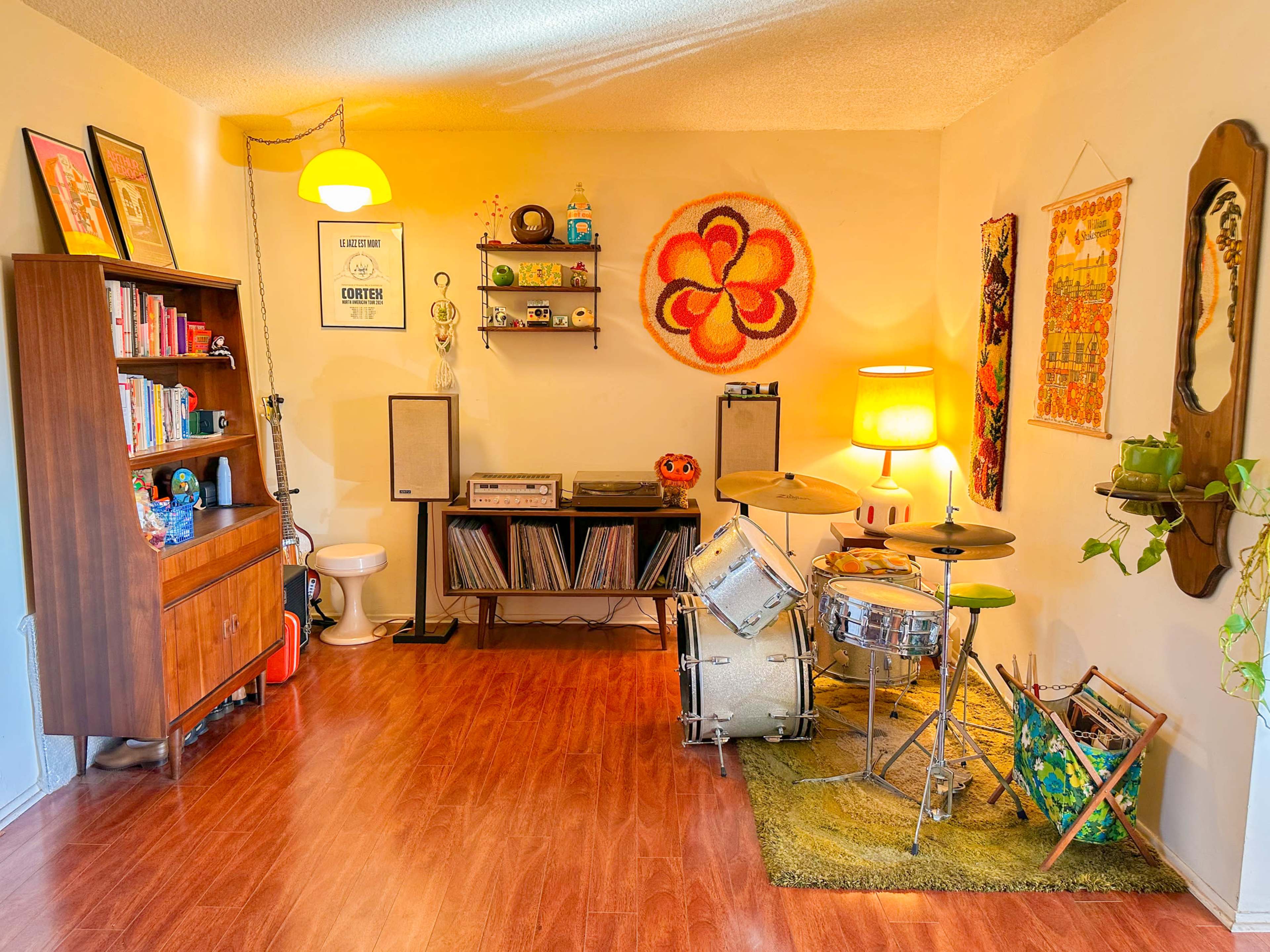 A cozy room features a drum set on a green rug, a wooden bookshelf filled with books, a record player, and colorful wall art.