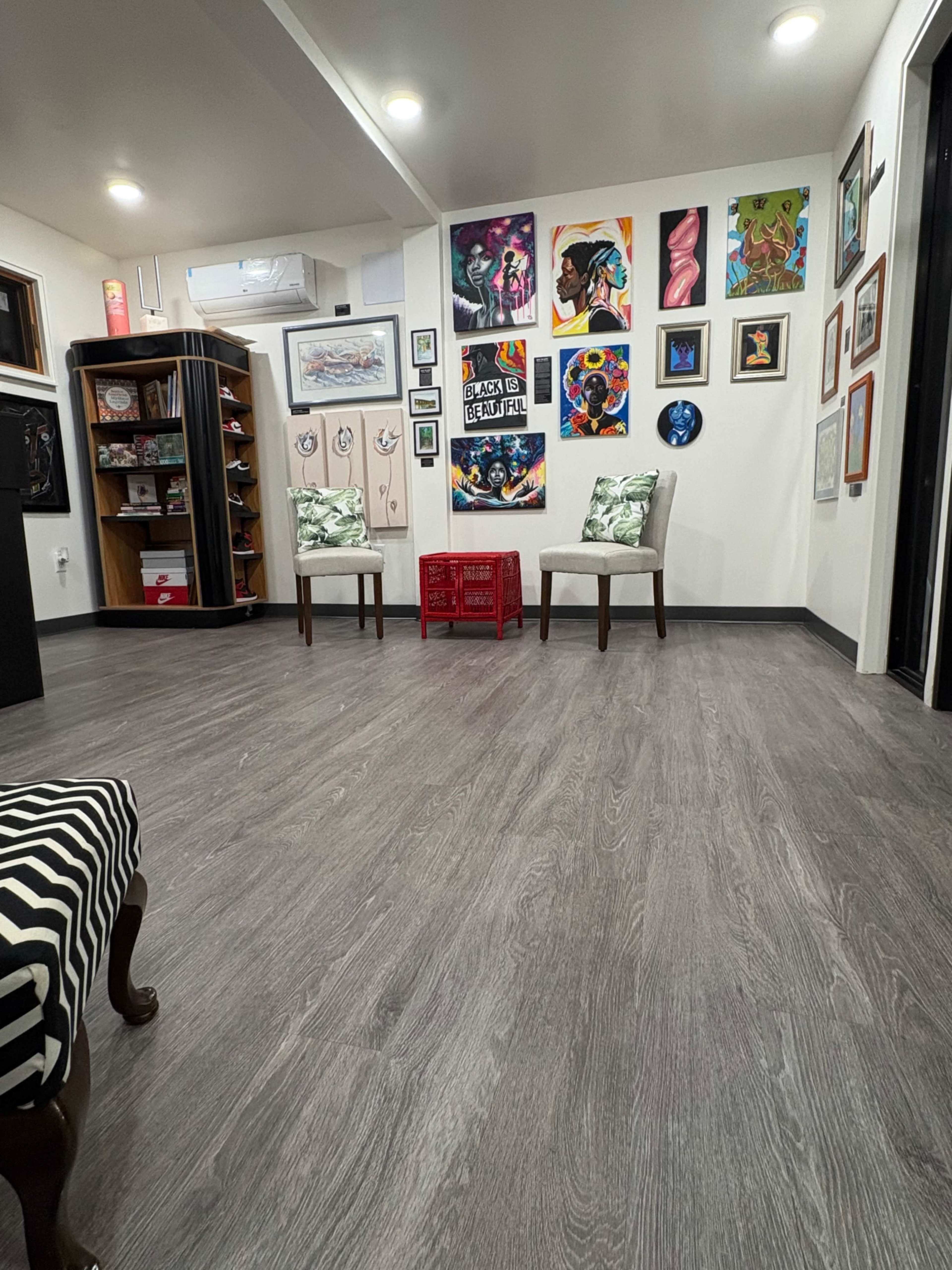 The image shows a modern indoor space featuring two chairs with cushions, a red storage crate, and a wall adorned with various colorful artworks.