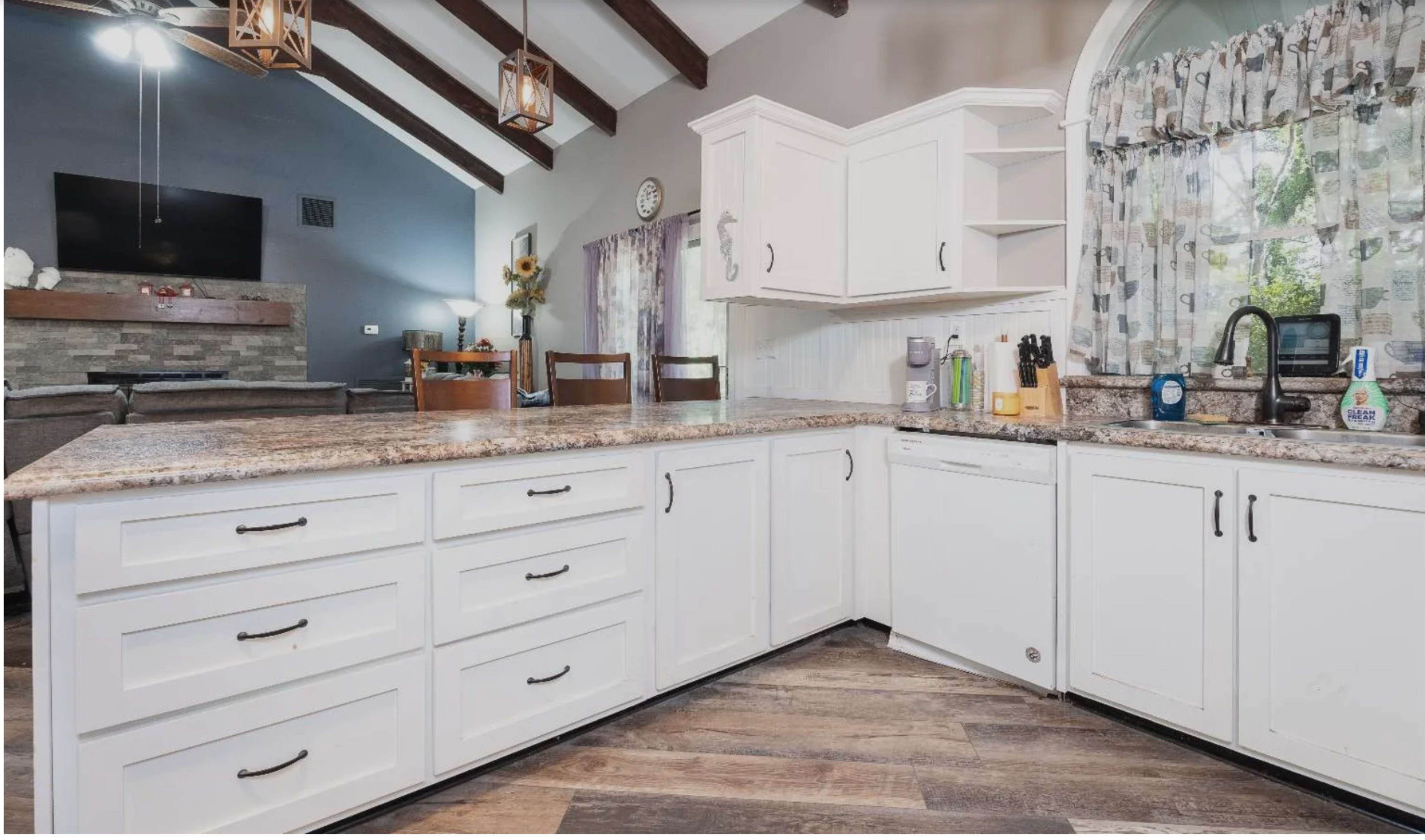 The image shows a modern kitchen with white cabinetry, a granite countertop, and a view of a living area in the background.