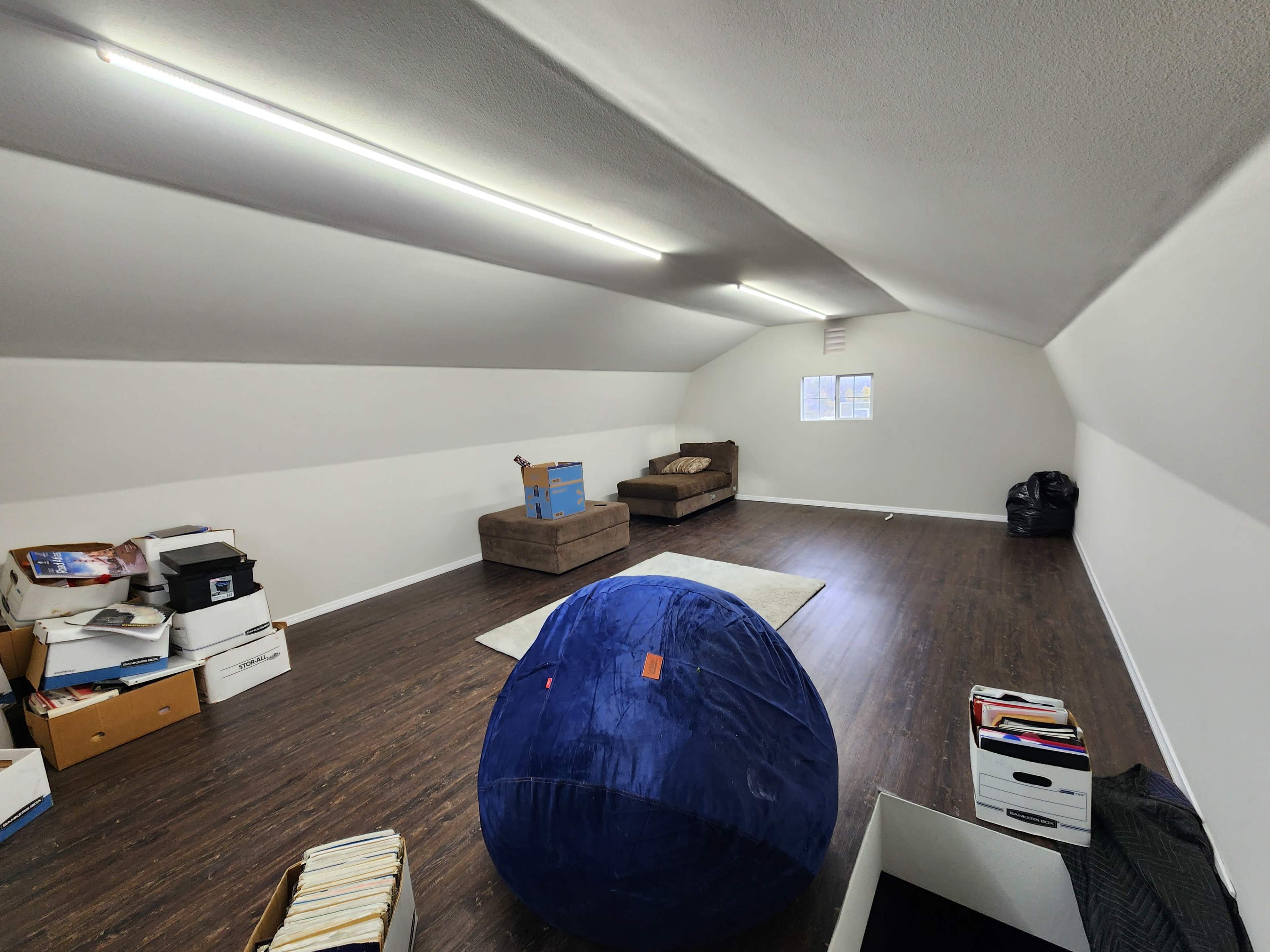 A spacious attic room with a wooden floor, a large blue bean bag, a brown couch, piles of boxes, and a small window.
