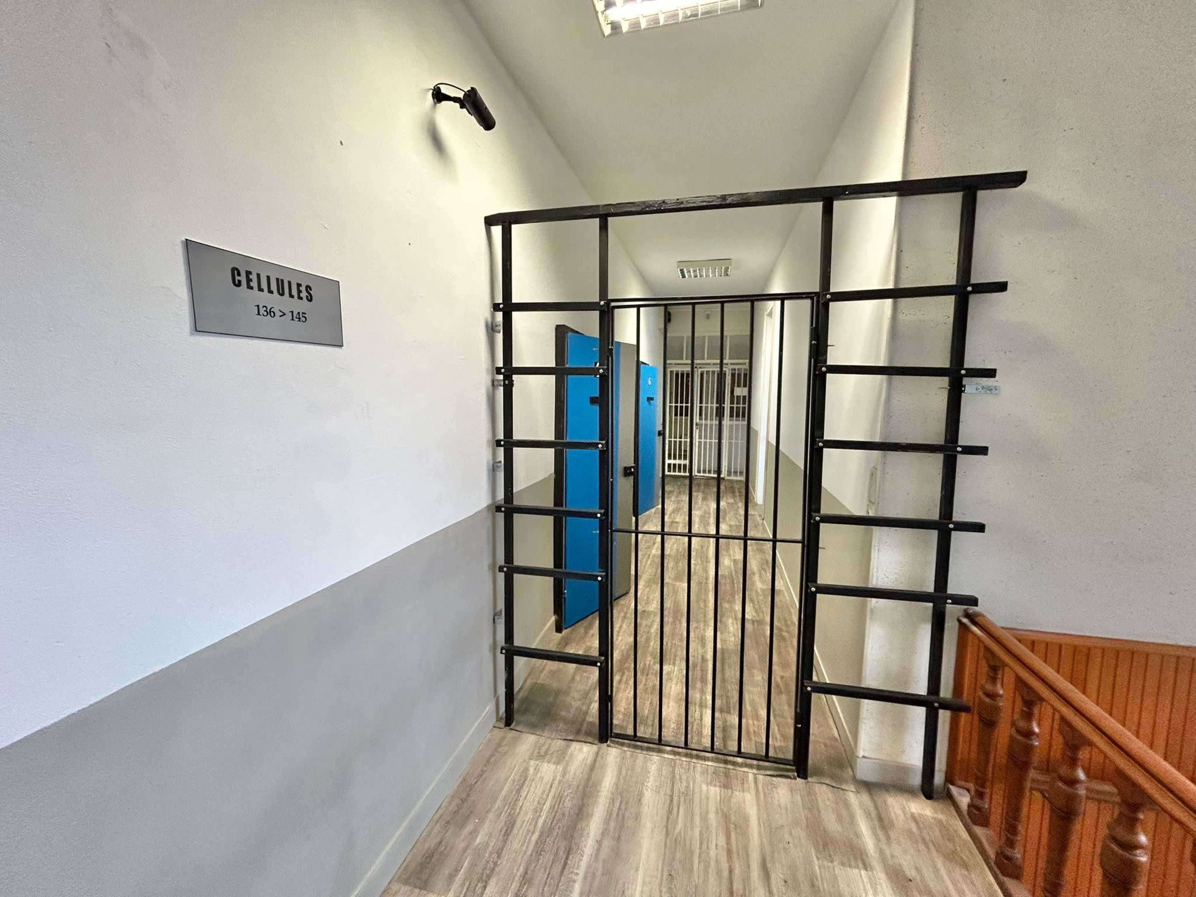 A corridor with a barred gate leading to a series of blue lockers and a cell at the end.