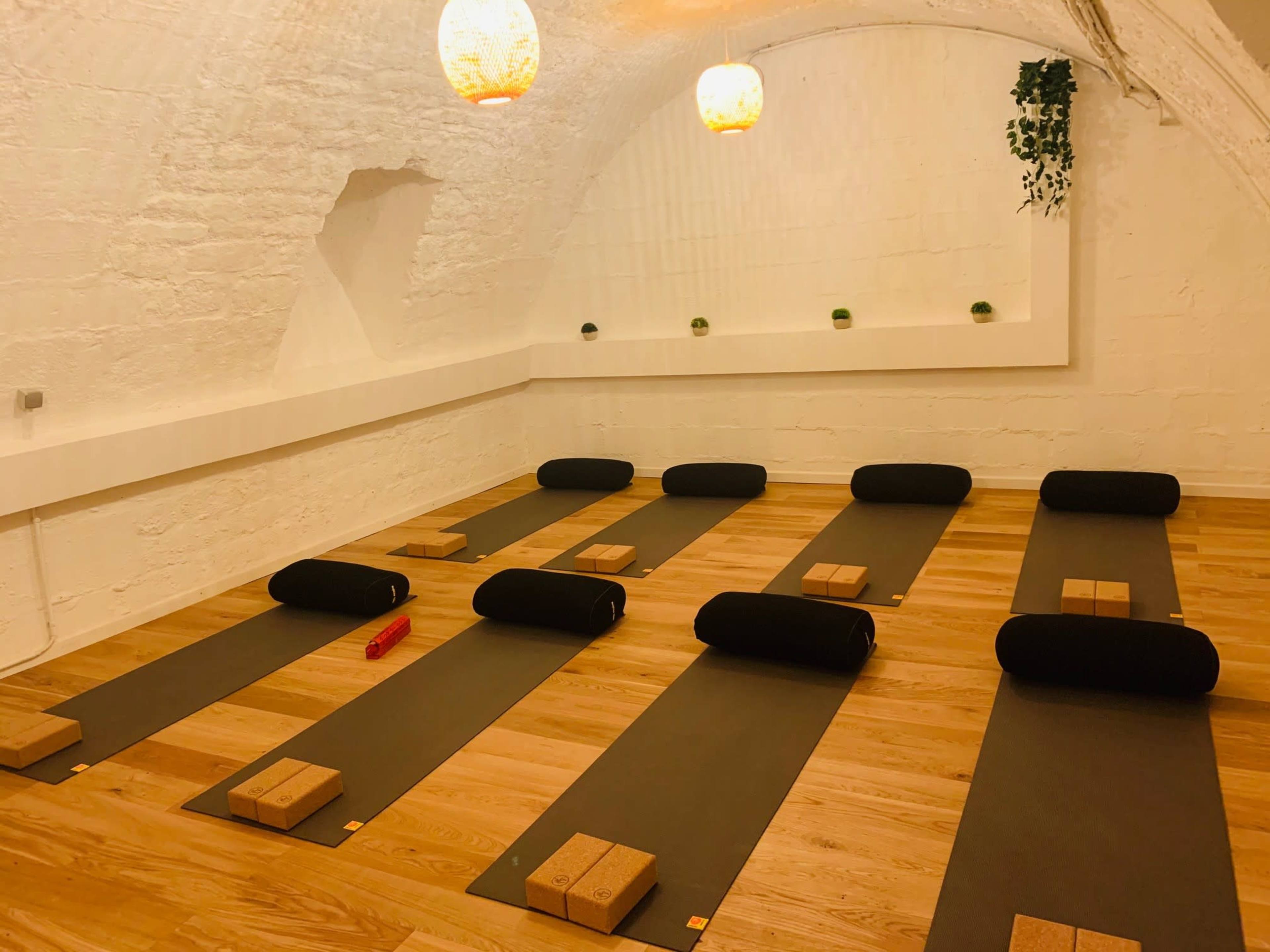 A yoga studio with several yoga mats arranged in neat rows, each accompanied by blocks and bolsters, under soft lighting.