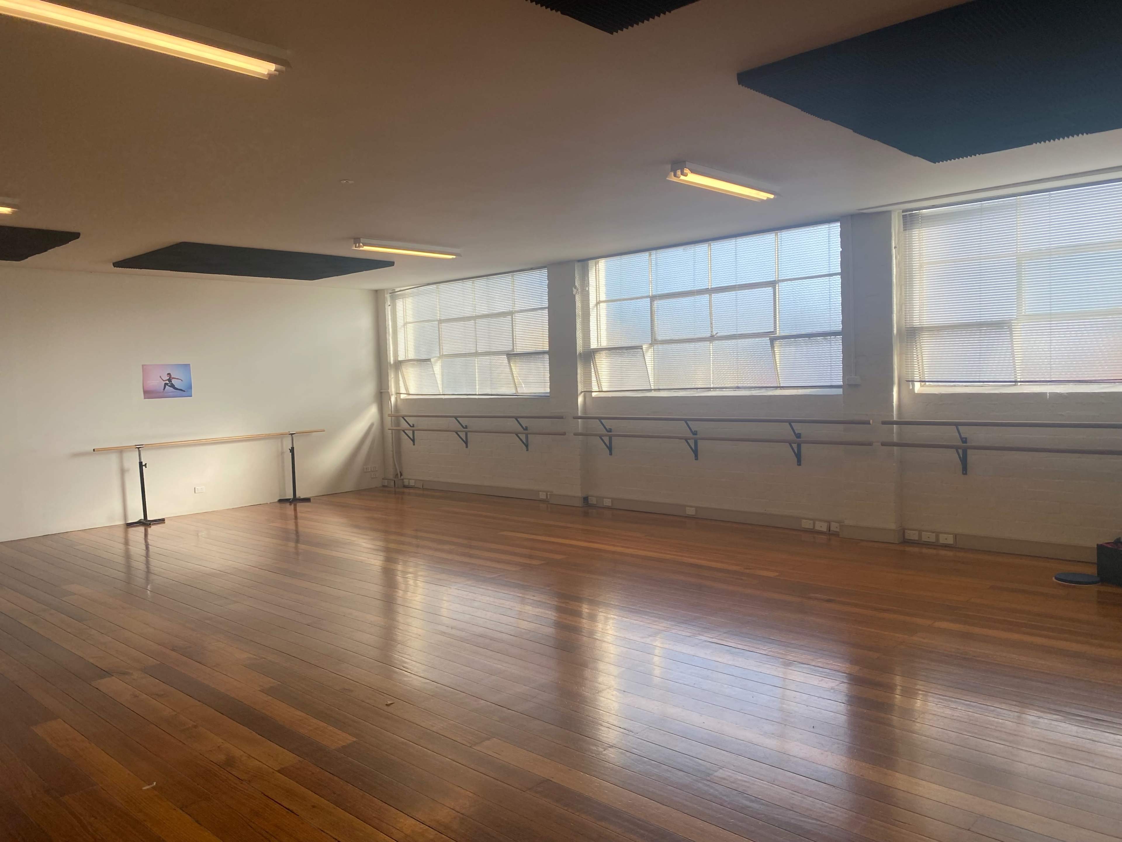 The image shows a spacious dance studio with wooden floors, large windows, and a ballet barre along the wall.