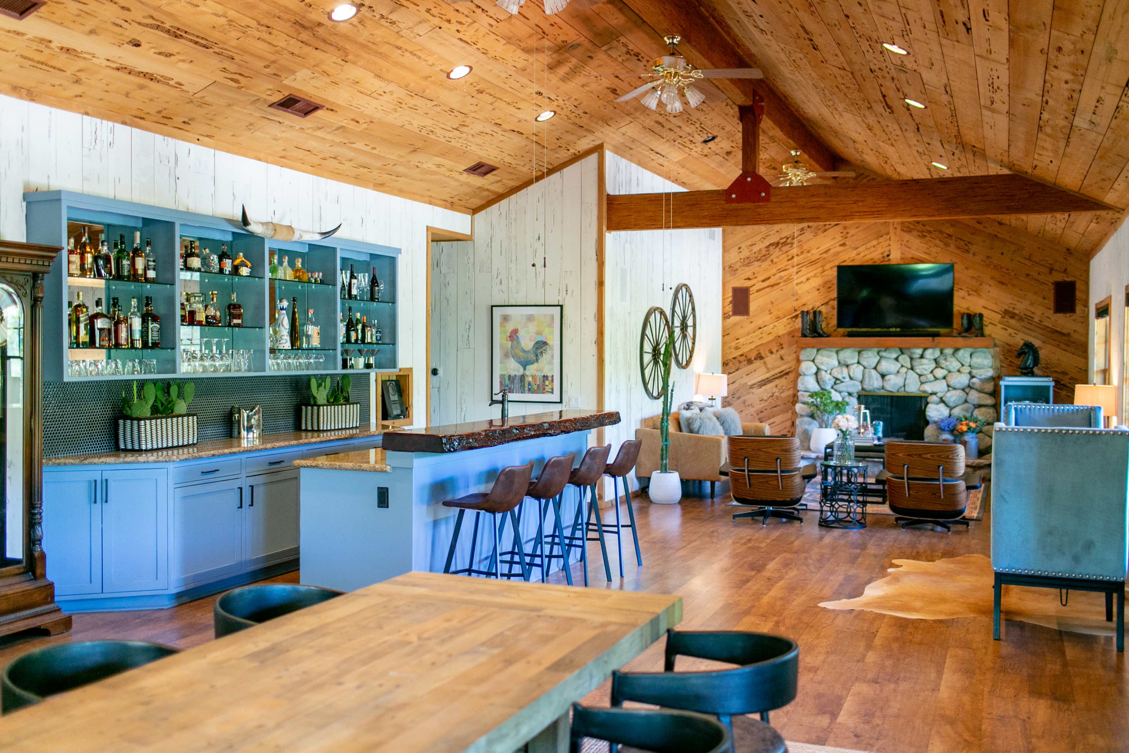 The image shows a spacious, modern living area with a bar, a stone fireplace, and large windows, featuring wooden accents throughout.