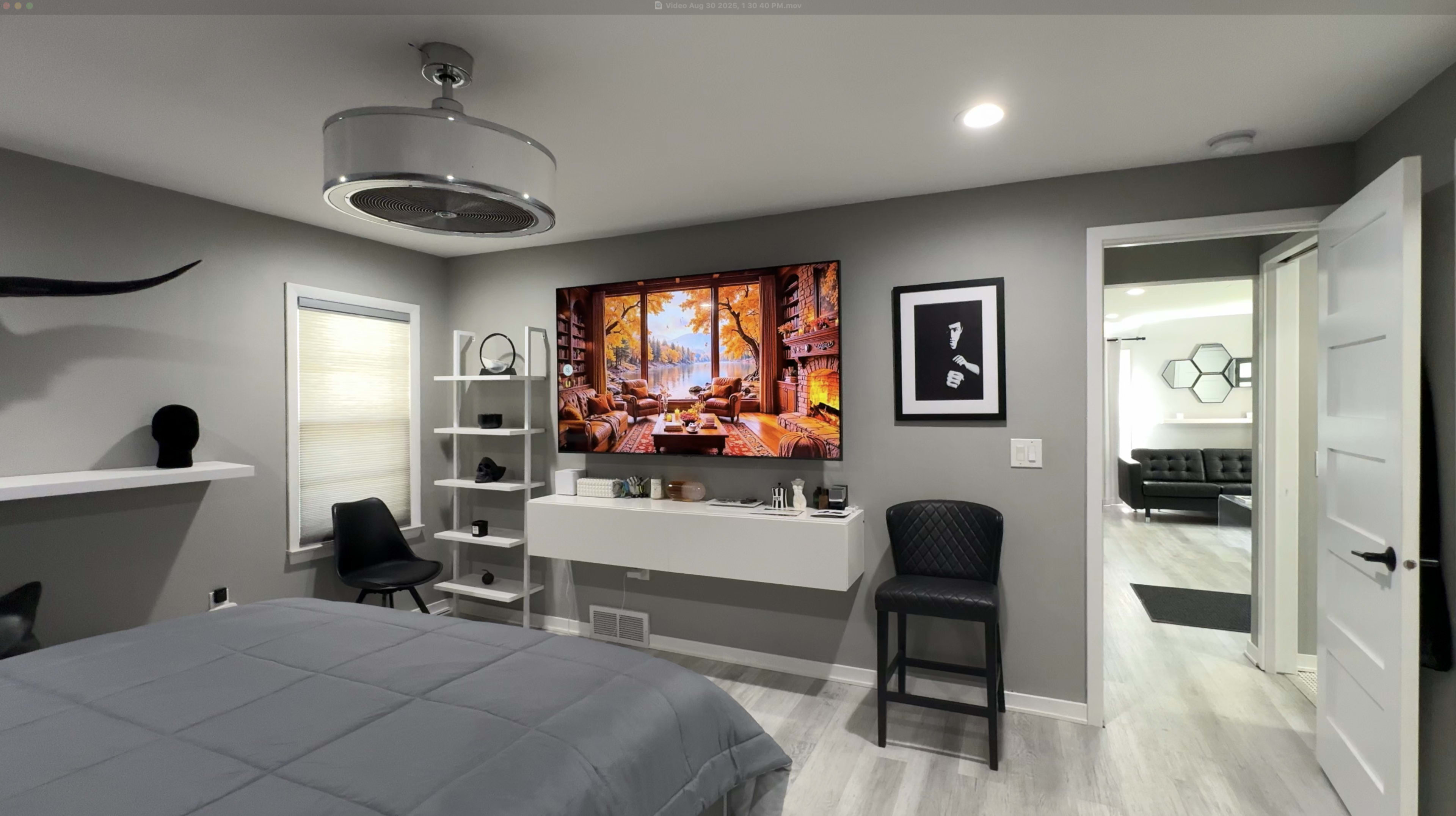 The image shows a modern bedroom with a wall-mounted television displaying a scenic landscape, a minimalist shelf, a chair, and doorways leading to another room and a closet.