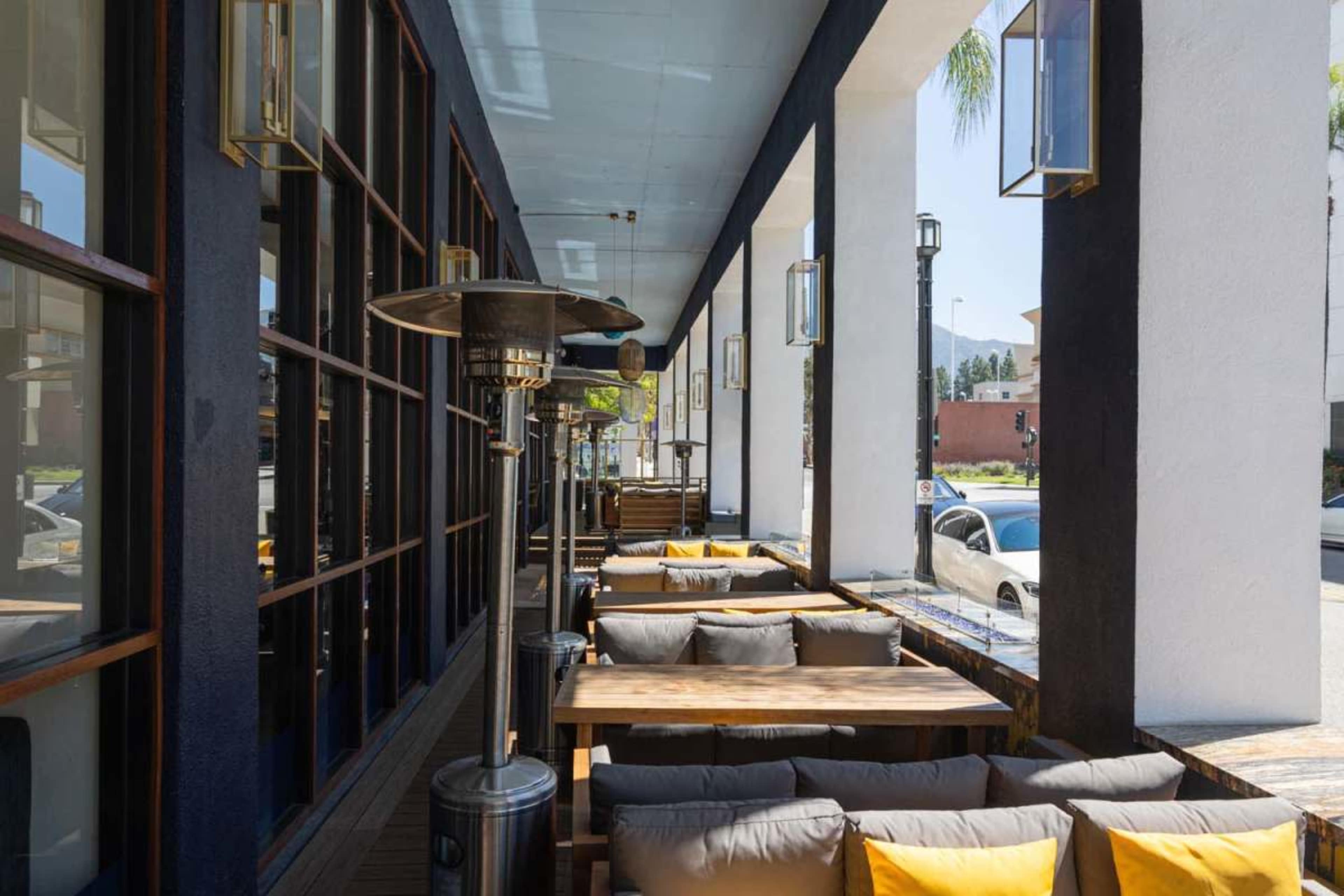 The image shows a row of outdoor seating with wooden tables and cushioned benches under a covered patio with large windows and heating lamps.