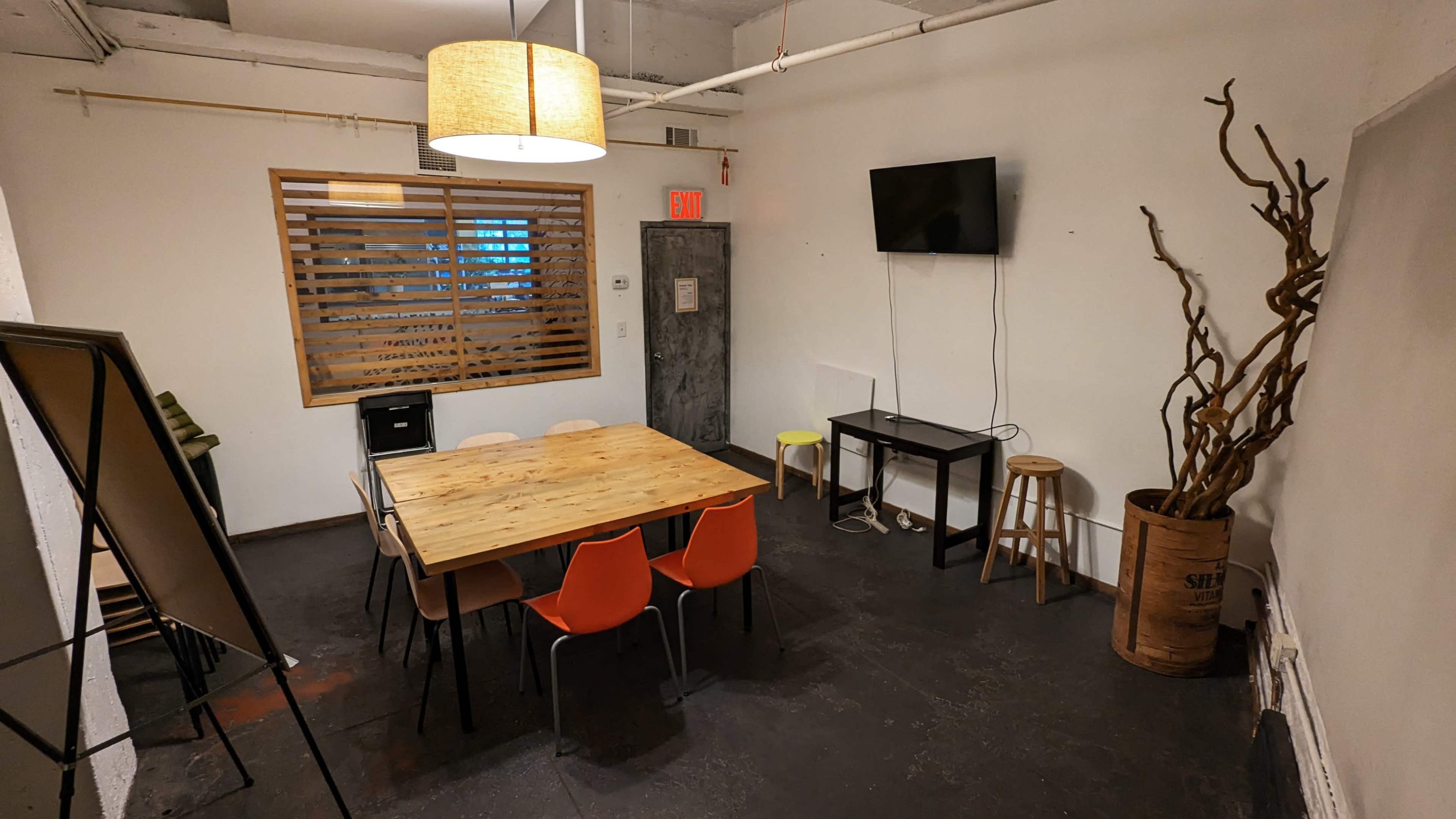 A small meeting room with a wooden table surrounded by colorful chairs, a wall-mounted television, and a decorative plant arrangement in a container.