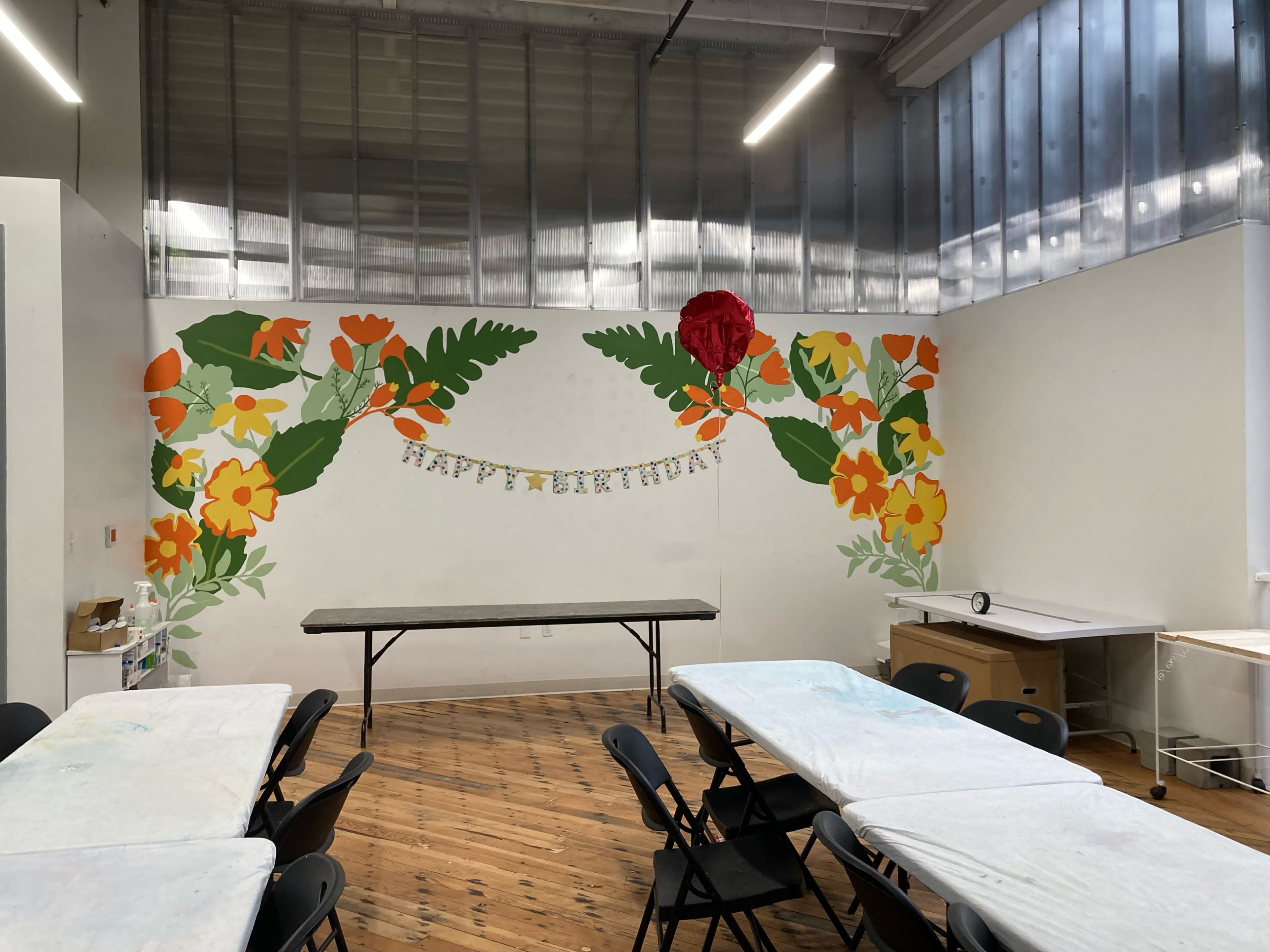 A brightly decorated room with a mural of flowers and leaves on one wall, a long table in the center, and folding chairs arranged around it.
