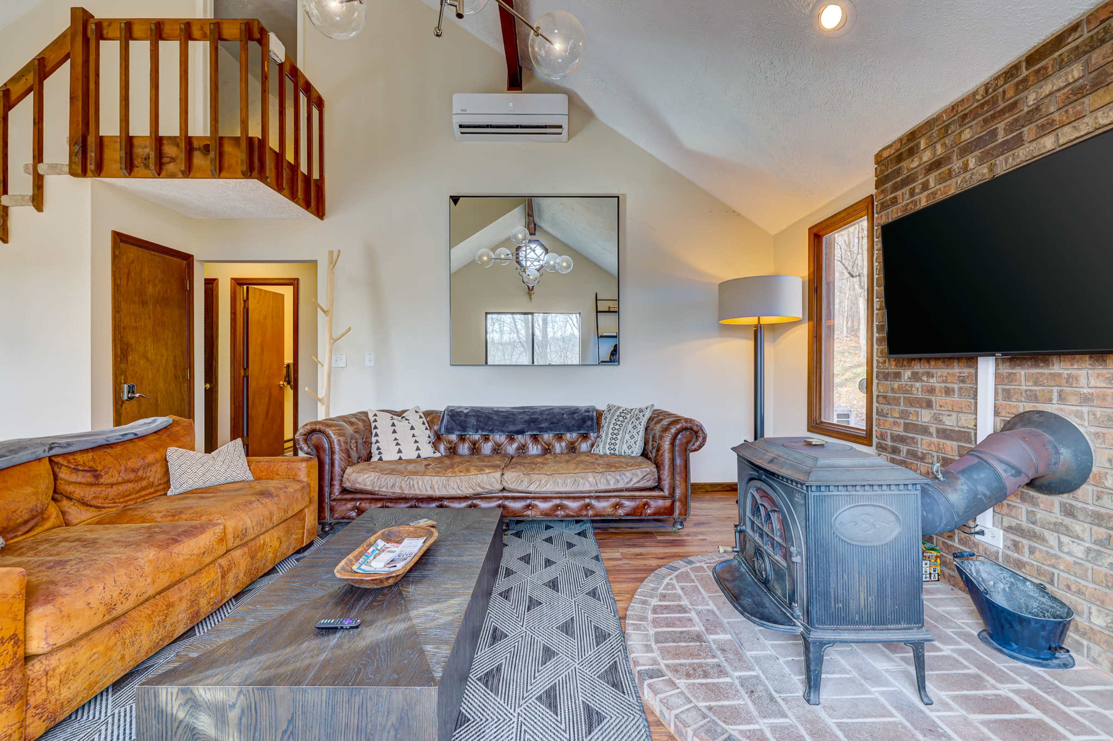 The image shows a cozy living room featuring a leather sofa, a wooden coffee table, a brick wall with a mounted TV, and a cast iron stove.