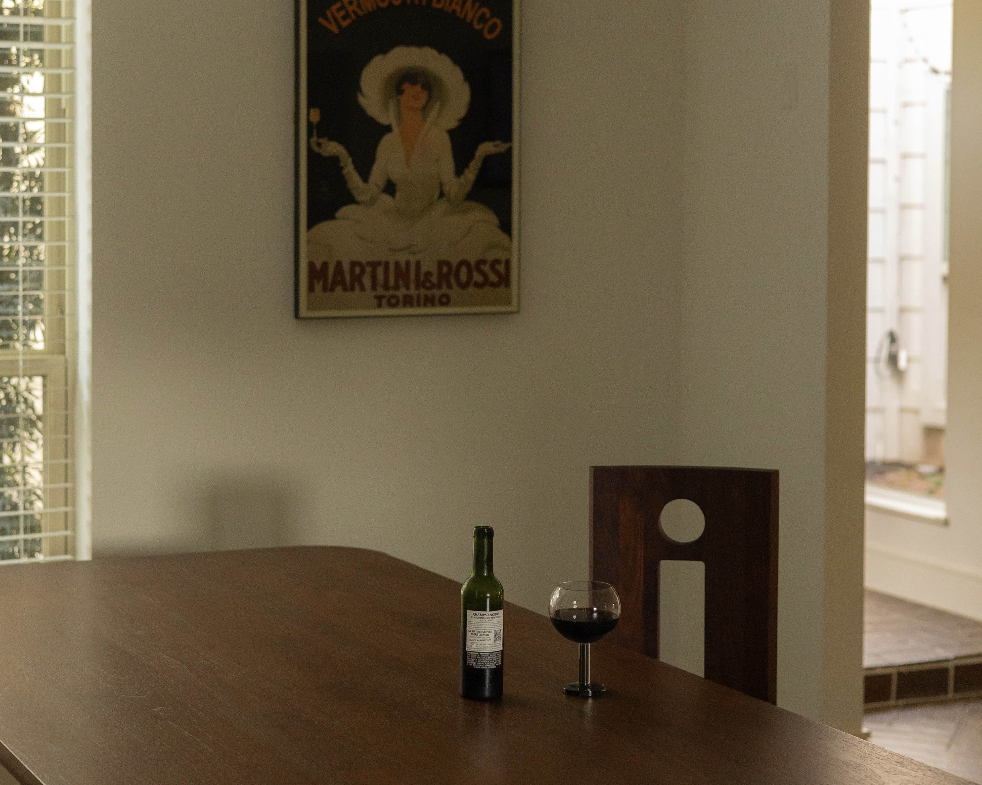 A wooden dining table sits in a room with a wine glass and a green bottle, next to a vintage poster on the wall.
