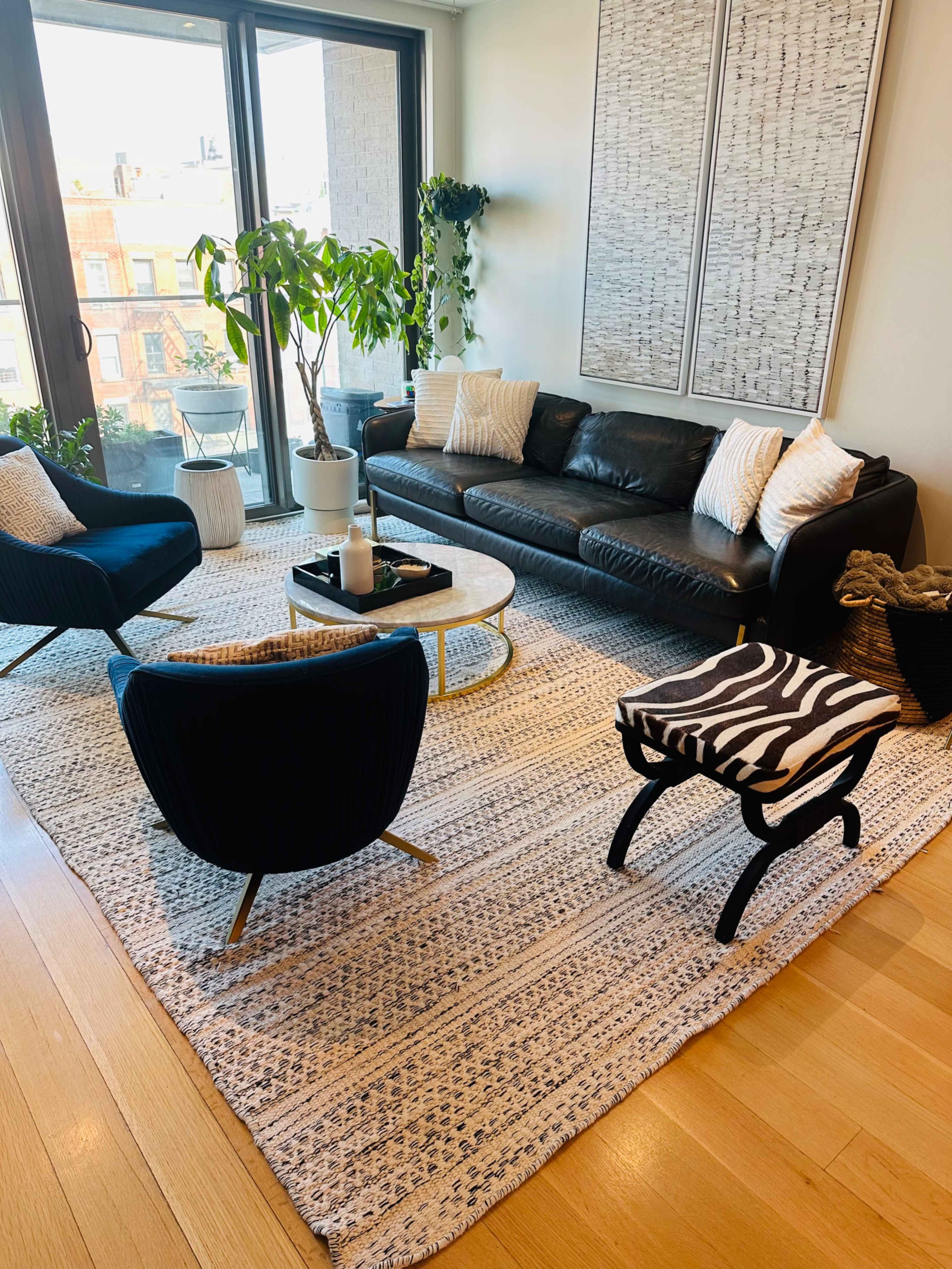 A modern living room with a black leather sofa, two black armchairs, a round coffee table, and a patterned rug, all arranged around a large window with plants nearby.