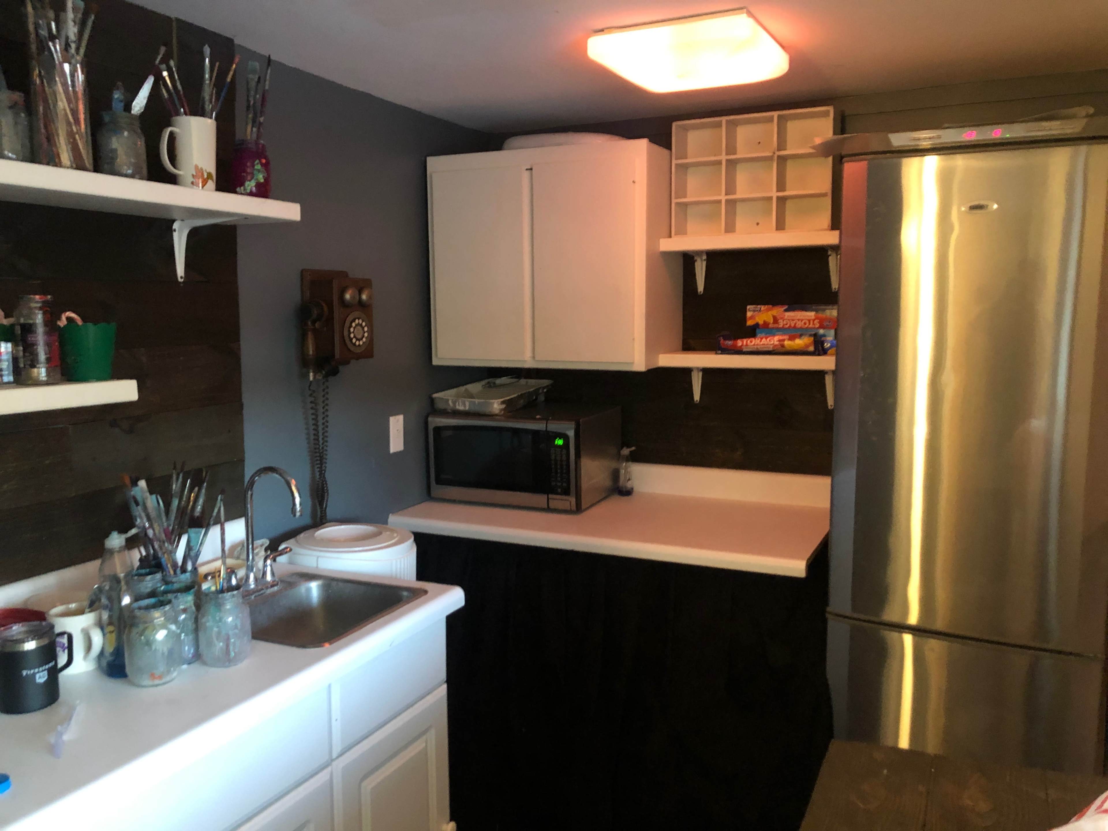A small kitchen area with a sink, microwave, refrigerator, and shelves holding various items and art supplies.