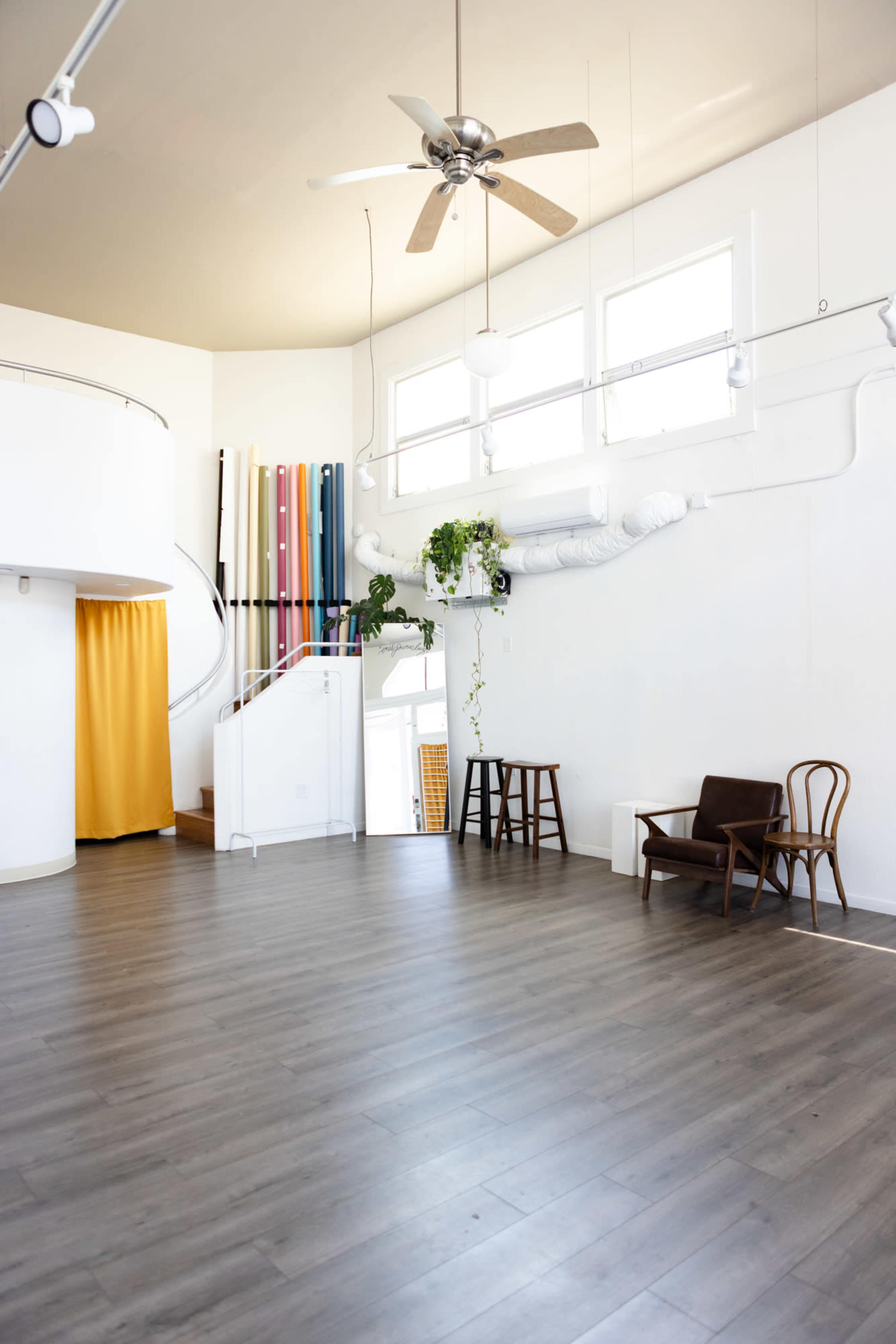 The image shows a spacious, well-lit room featuring a wooden floor, a spiral staircase, and colorful vertical wall accents.