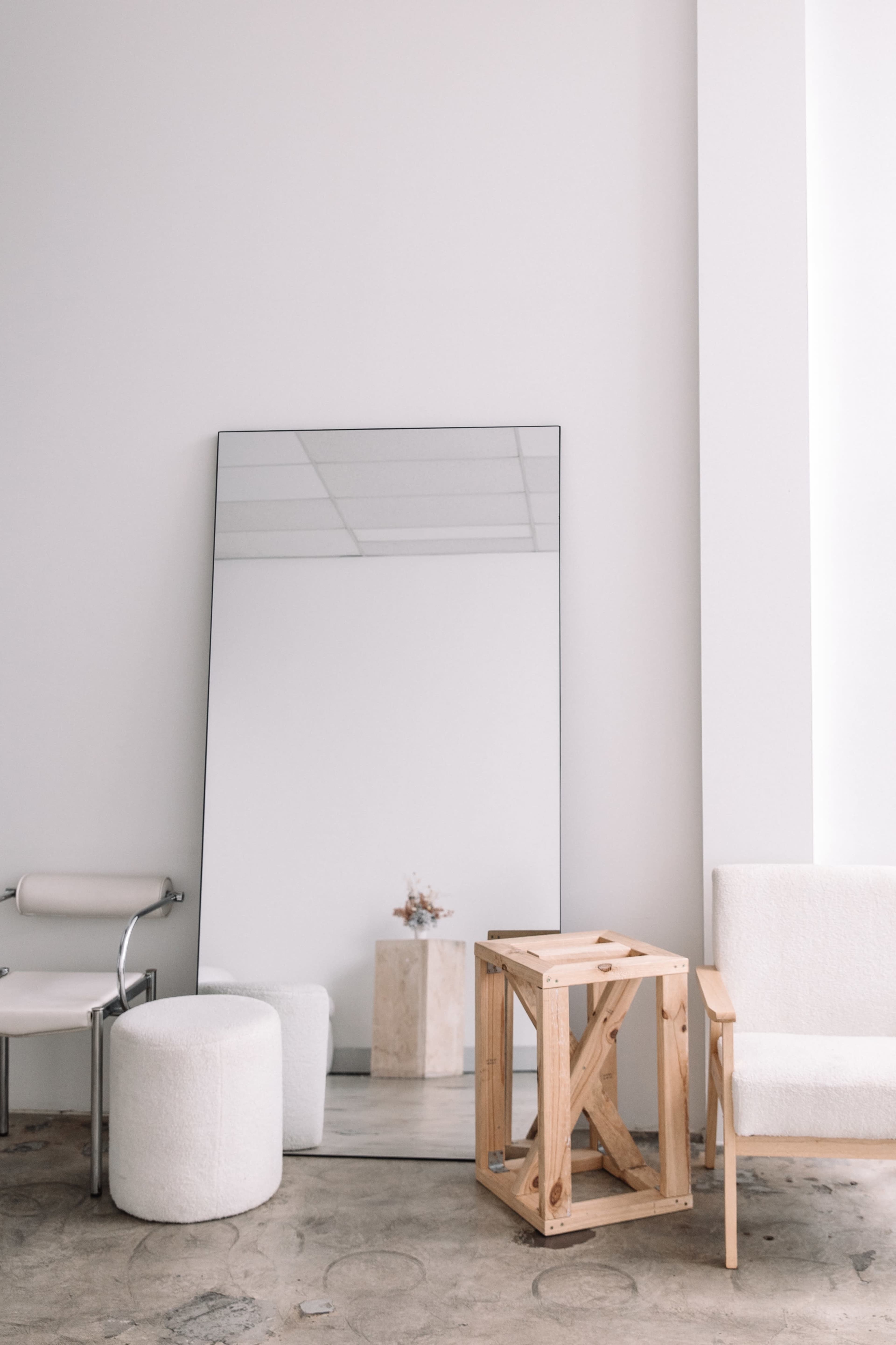 A large mirror leans against a plain wall in a minimalist room featuring a wooden crate, a cylindrical ottoman, and a chair.