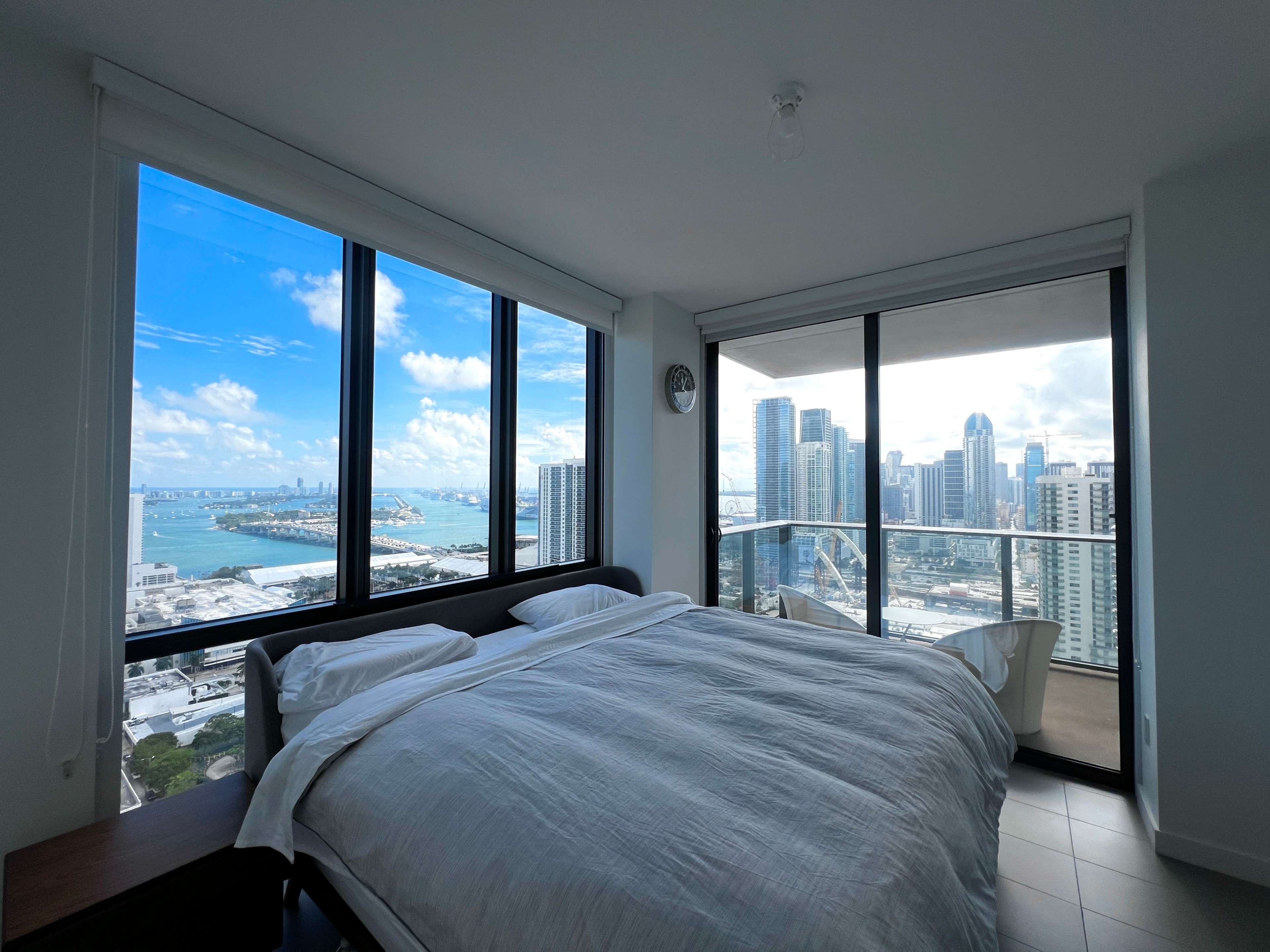 A modern bedroom features a large window with a city and ocean view, showcasing a skyline with tall buildings against a blue sky.