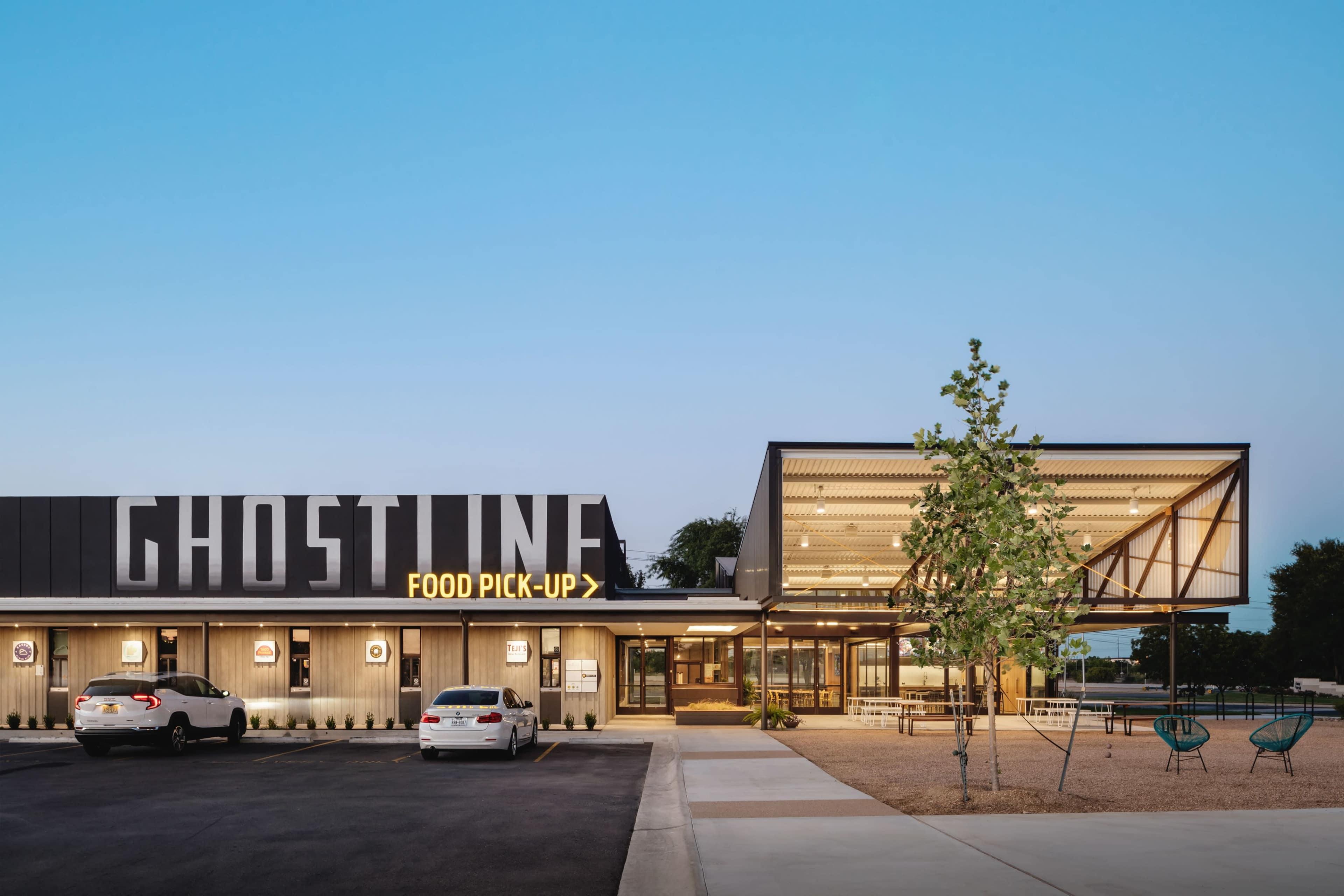 The image shows a modern building with a prominent sign reading "GHOSTLINE" and a food pickup area, featuring a landscaped exterior and a clear blue sky.