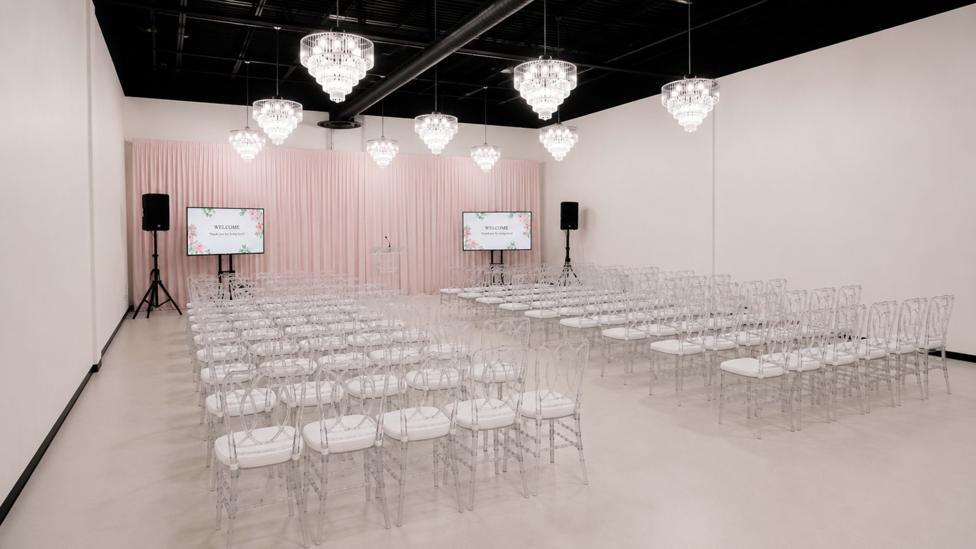The image shows an elegantly arranged event space with rows of clear chairs, a podium at the front, and digital screens displaying a welcome message, all set against a backdrop of soft pink drapery and chandeliers.