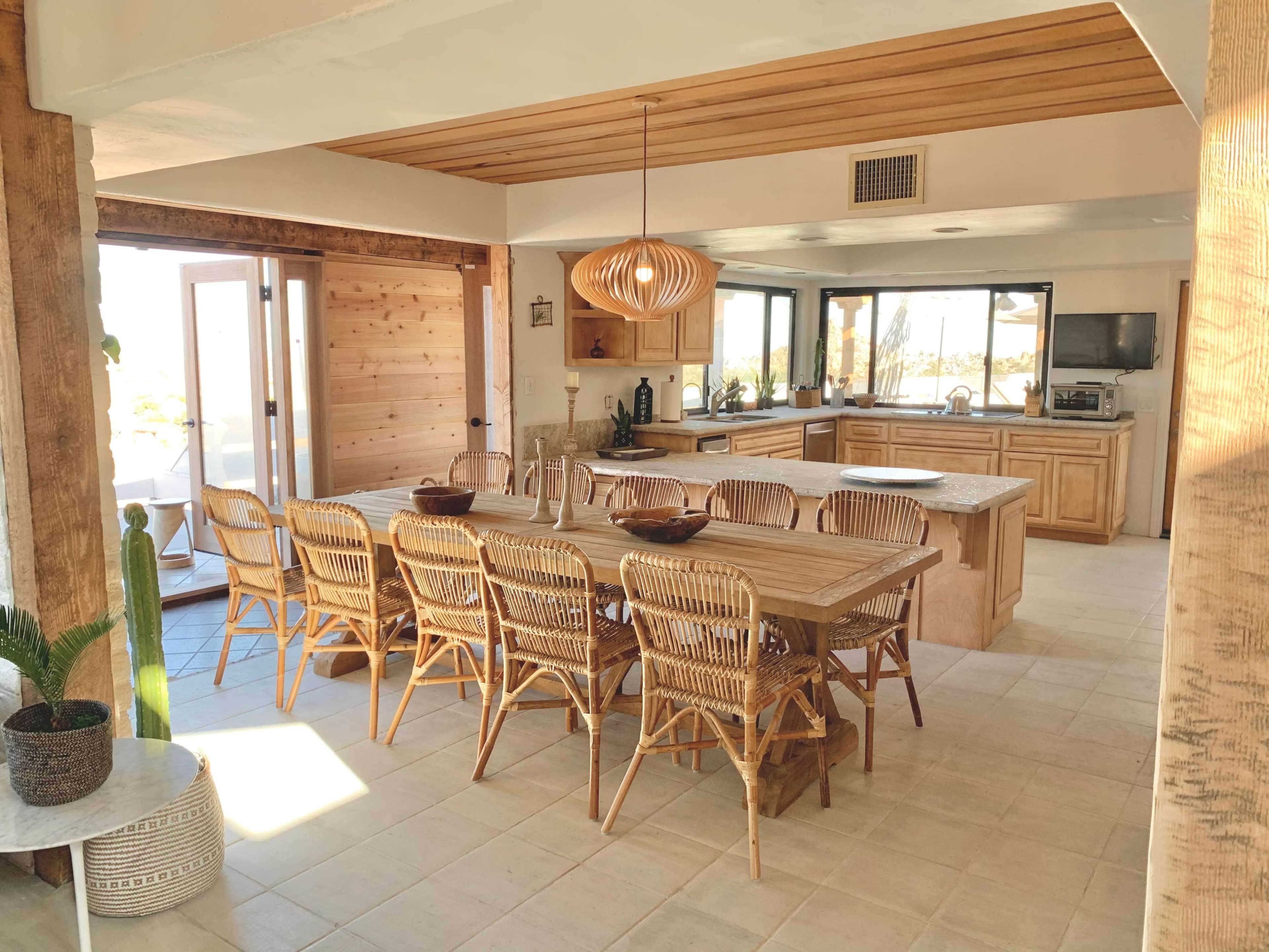A spacious kitchen and dining area features a large wooden table surrounded by rattan chairs, with light streaming in through multiple windows.