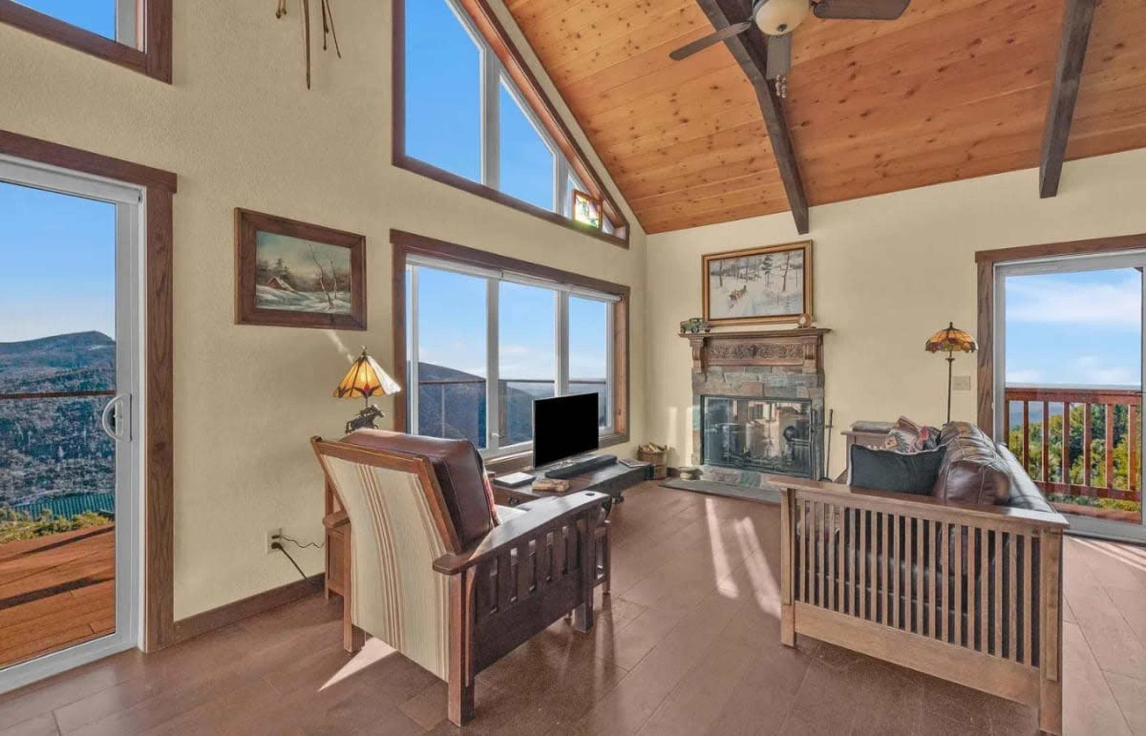 The image shows a living room with large windows, a fireplace, and a wooden ceiling, featuring a couch and an armchair near a television.