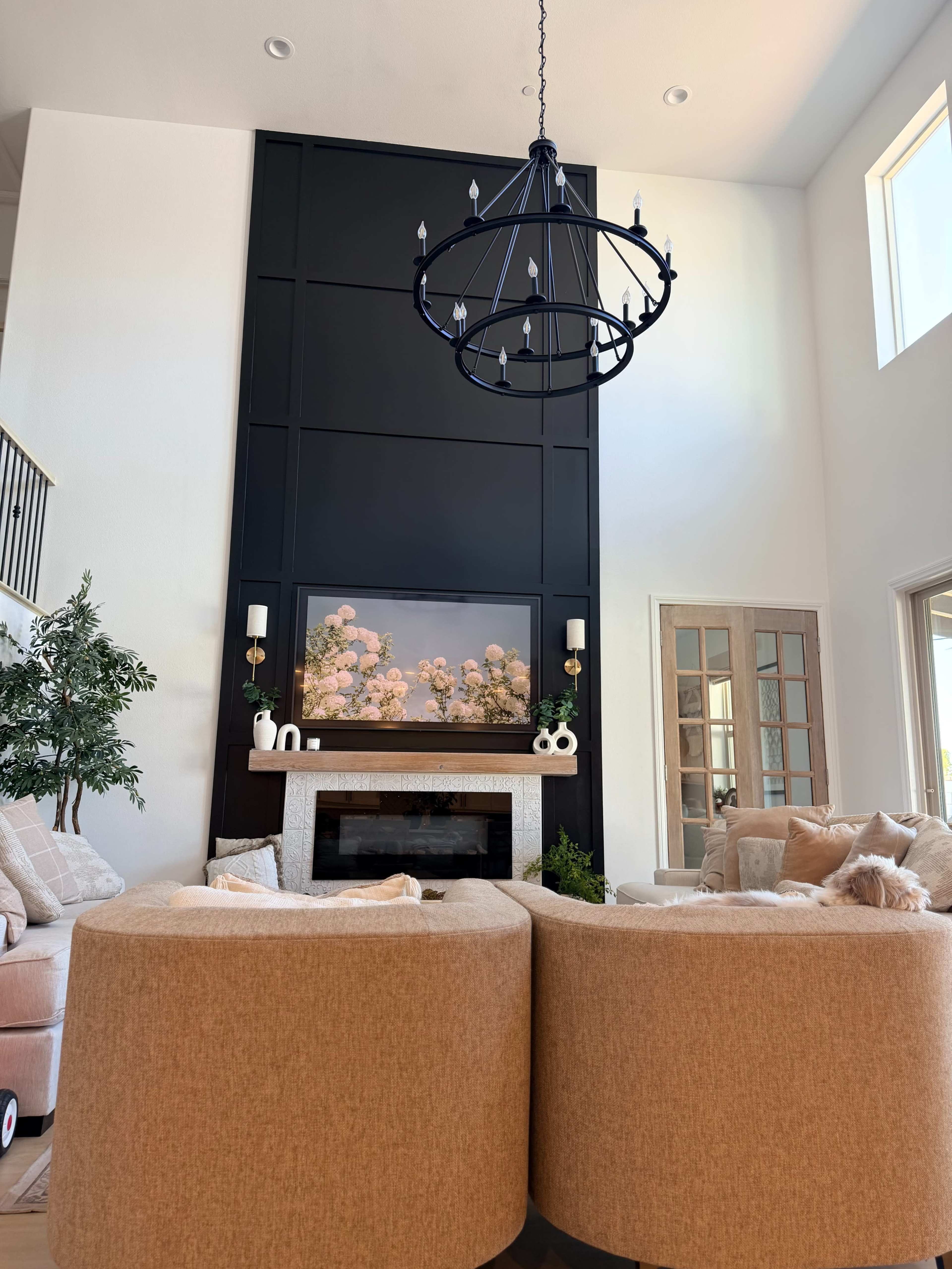 The image shows a modern living room with a large chandelier, a fireplace adorned with floral decor, and two rounded sofas facing each other.