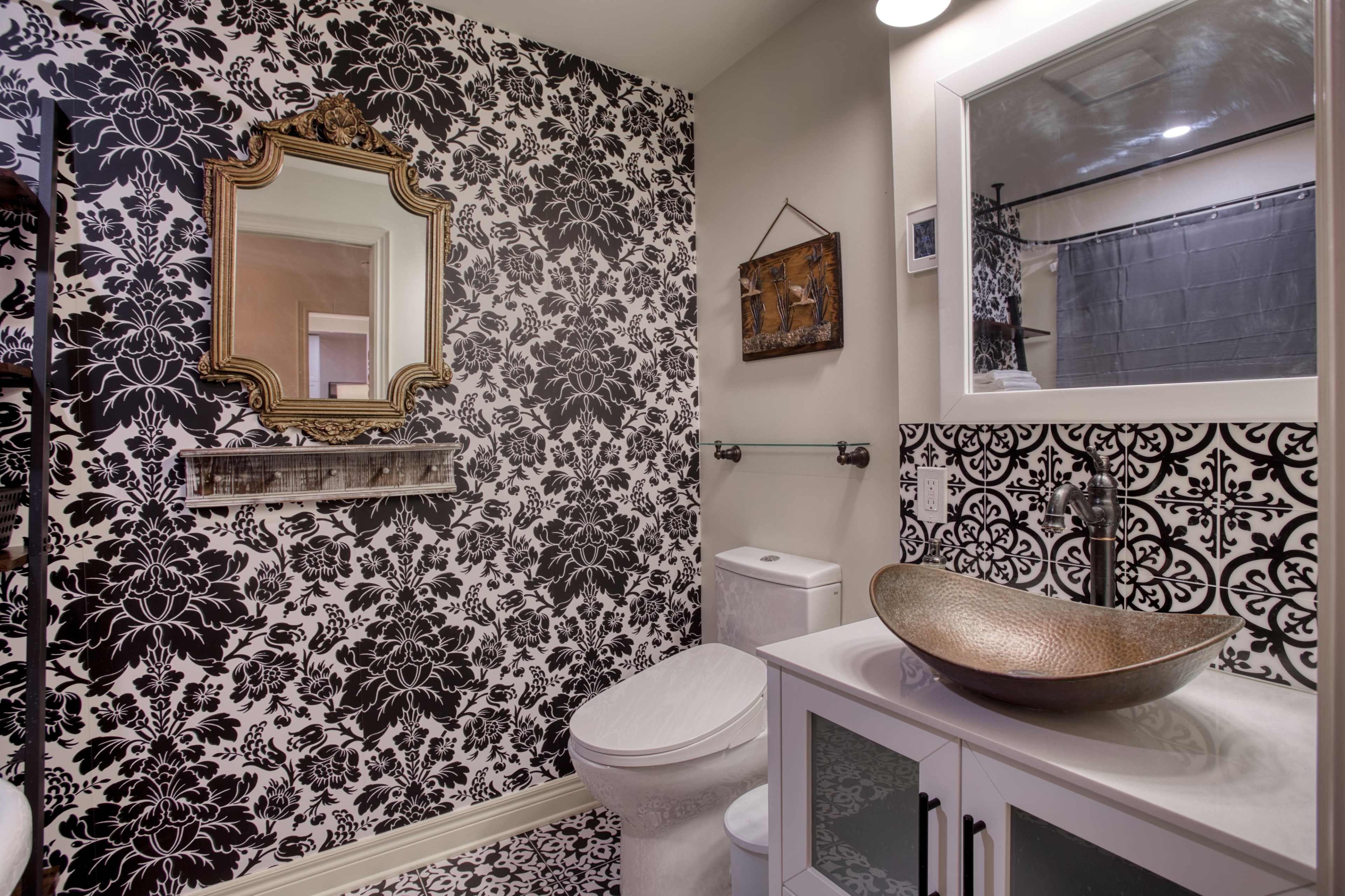 The image shows a bathroom with bold black and white floral wallpaper, a round decorative mirror, a modern sink with a bronze bowl, and a toilet.