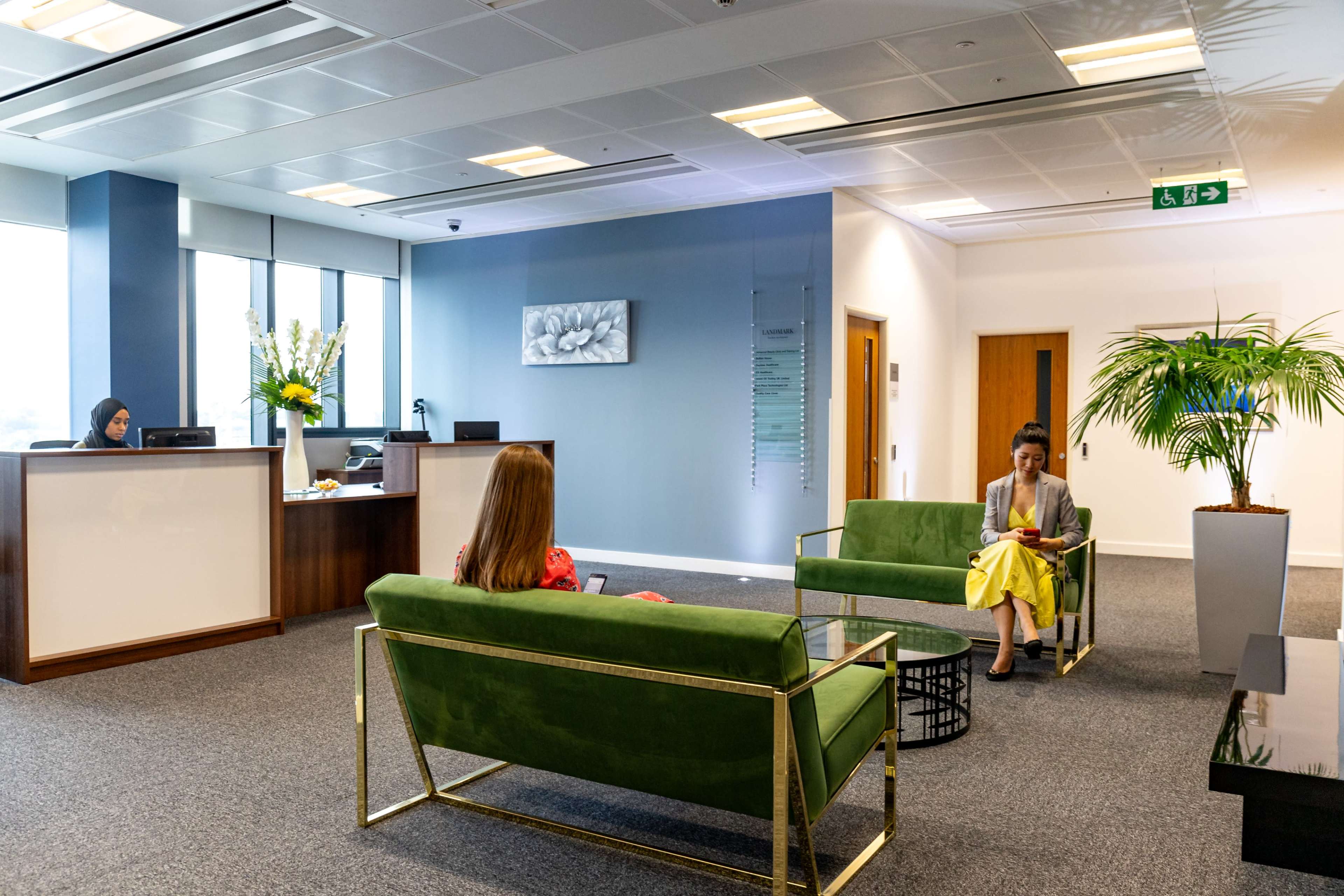 A modern office reception area with two women seated on green sofas, a reception desk in the background, and decorative plants.