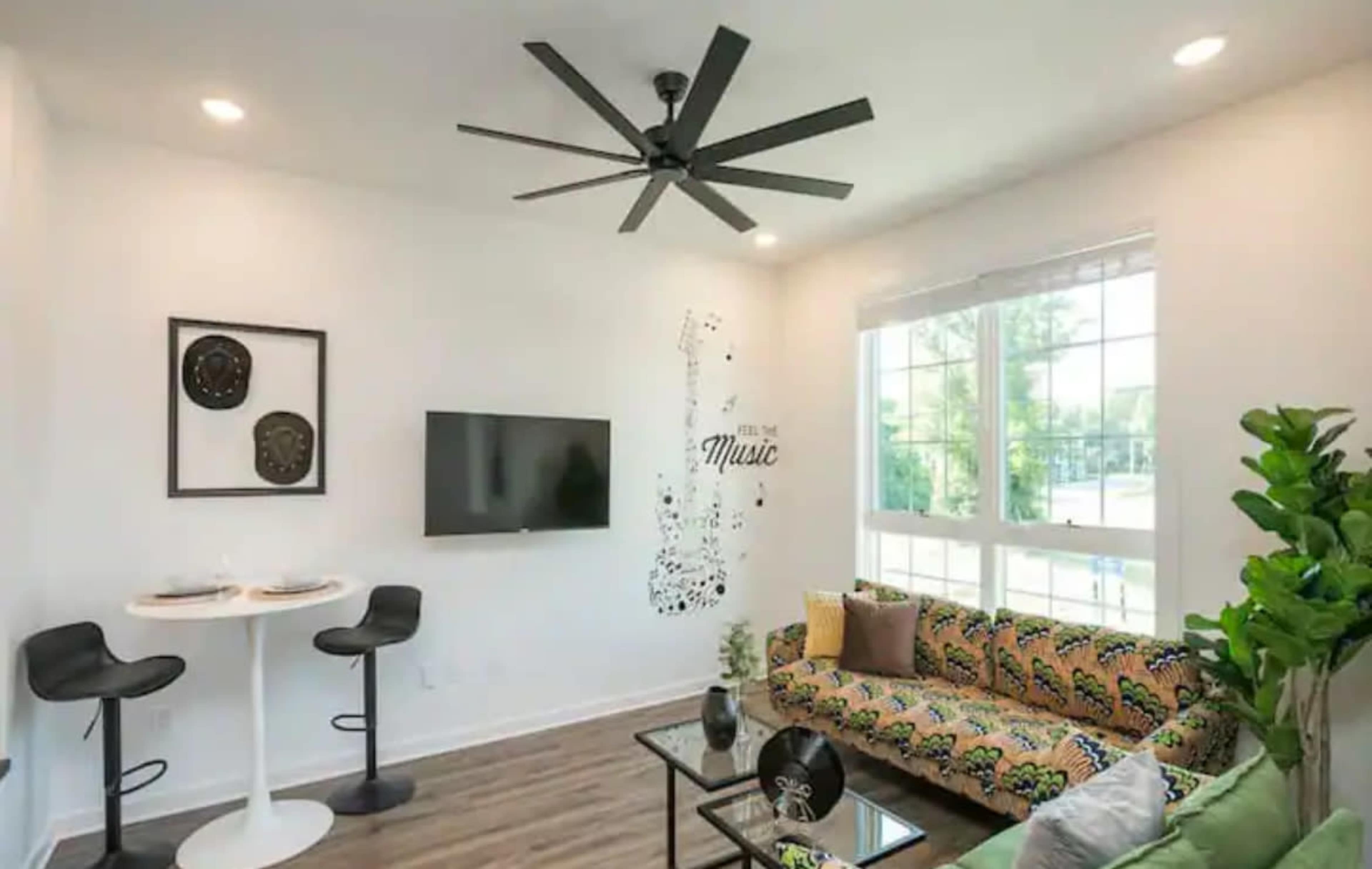 A modern living area with a green patterned sofa, a round white dining table with two stools, a wall-mounted TV, and a decorative wall design featuring musical notes.