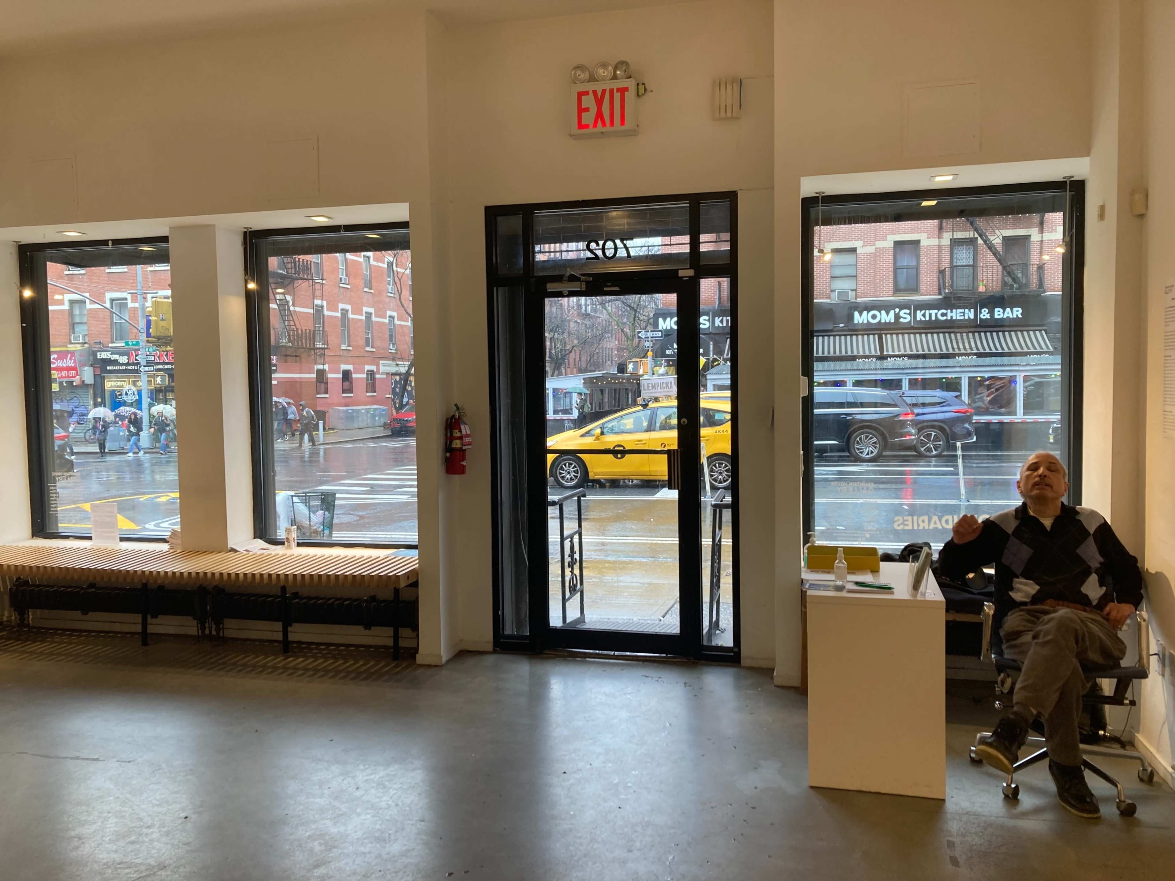 A man sits in a chair inside a minimalistic space, while large windows showcase a rainy street with a yellow taxi and pedestrians outside.