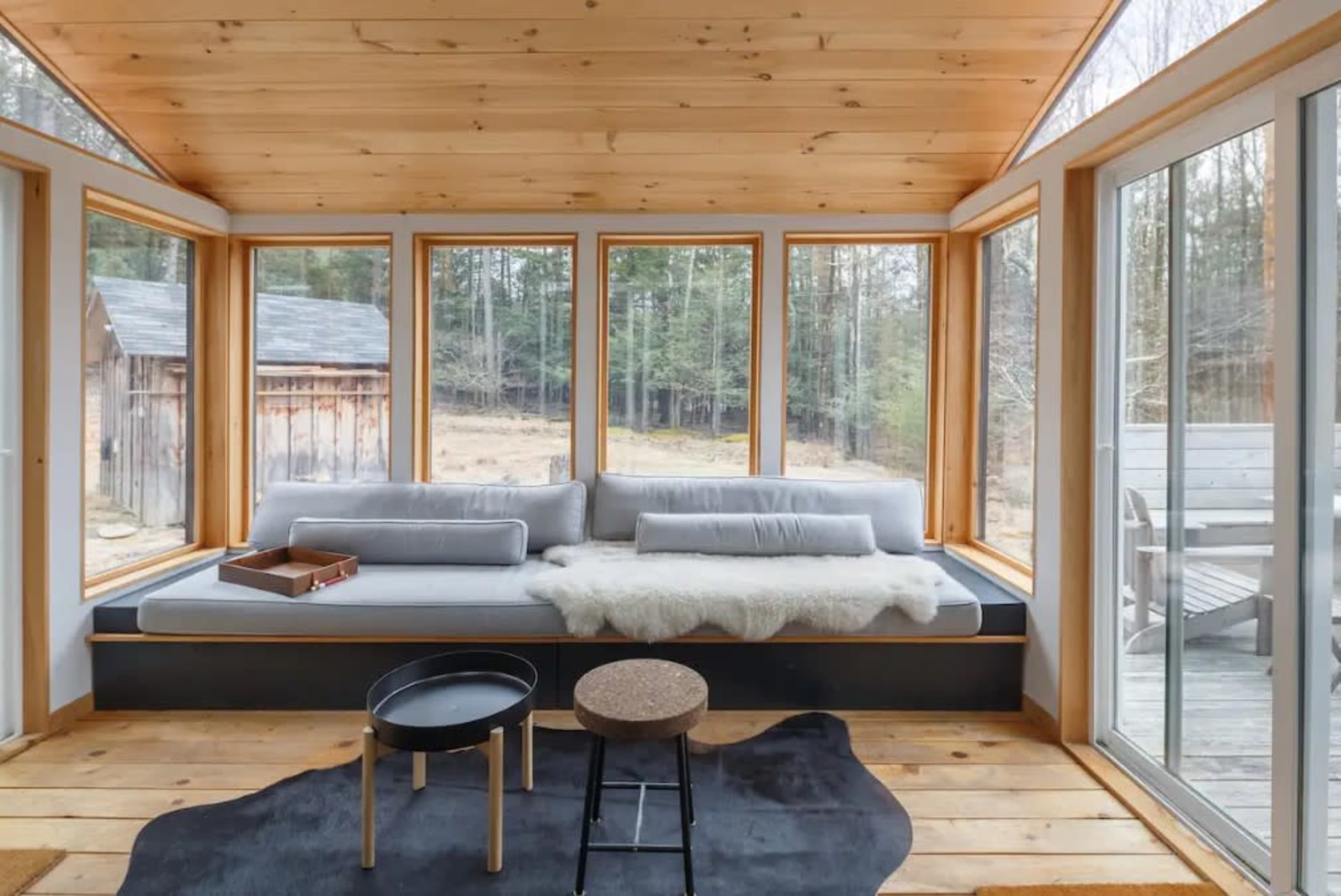 A sunlit room with large windows features a gray sofa, a wooden floor, and simple furniture including a round table and a stool.