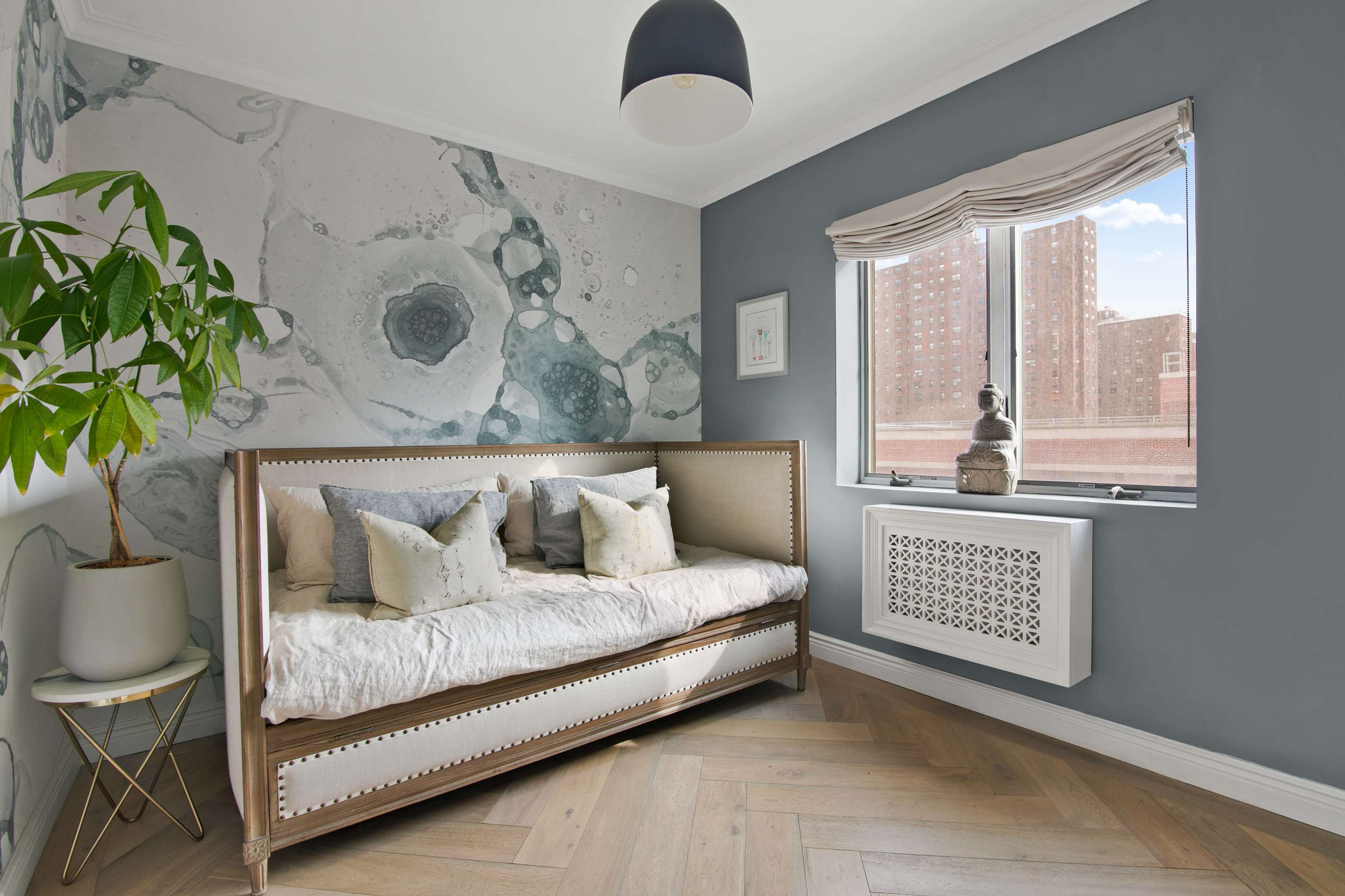 The image shows a stylish bedroom featuring a daybed with decorative pillows, a large window with city views, and a plant beside a gold side table.