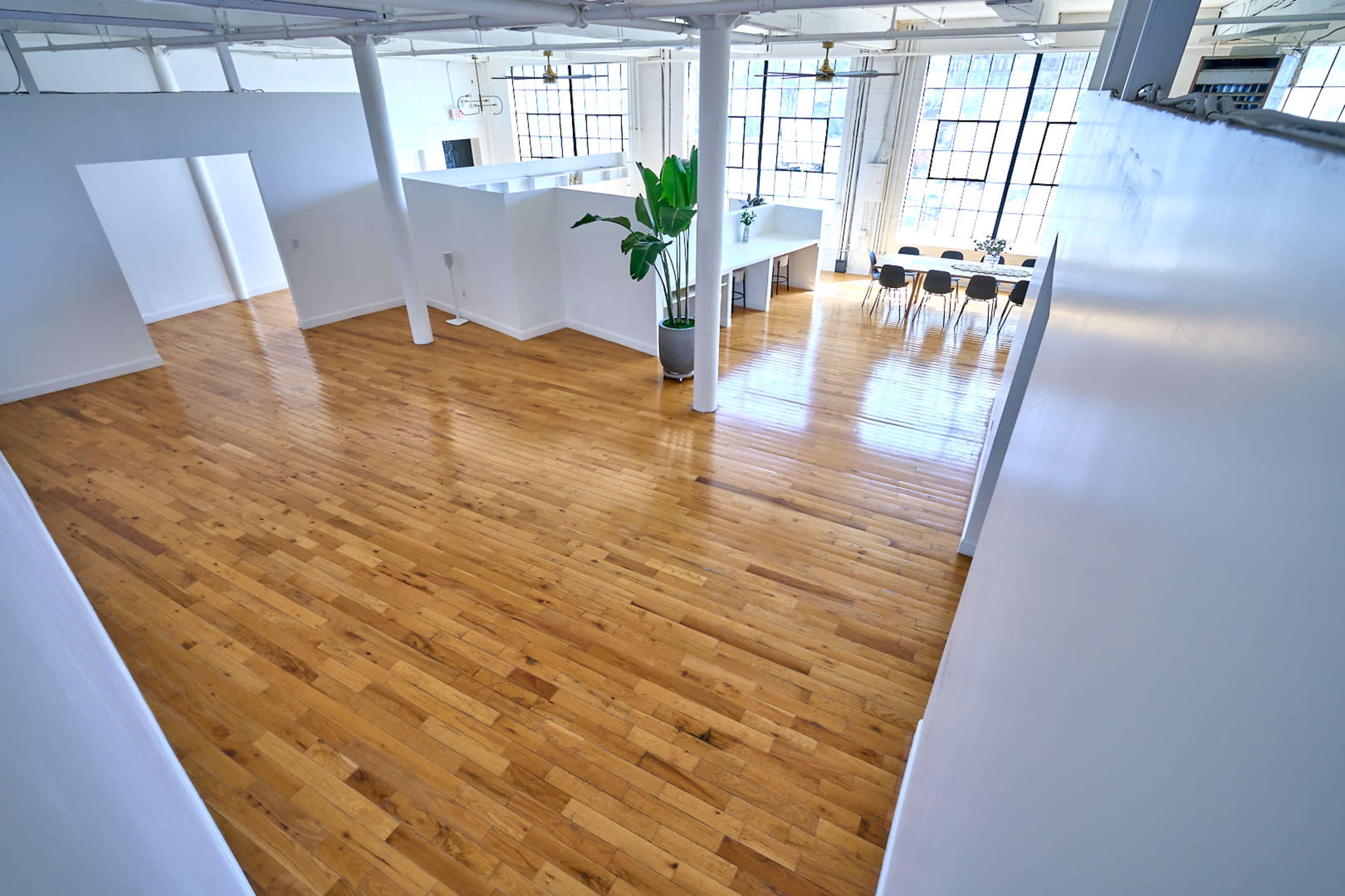 A spacious, well-lit interior with polished wooden floors, white walls, and large windows, featuring a dining area with a table and chairs.