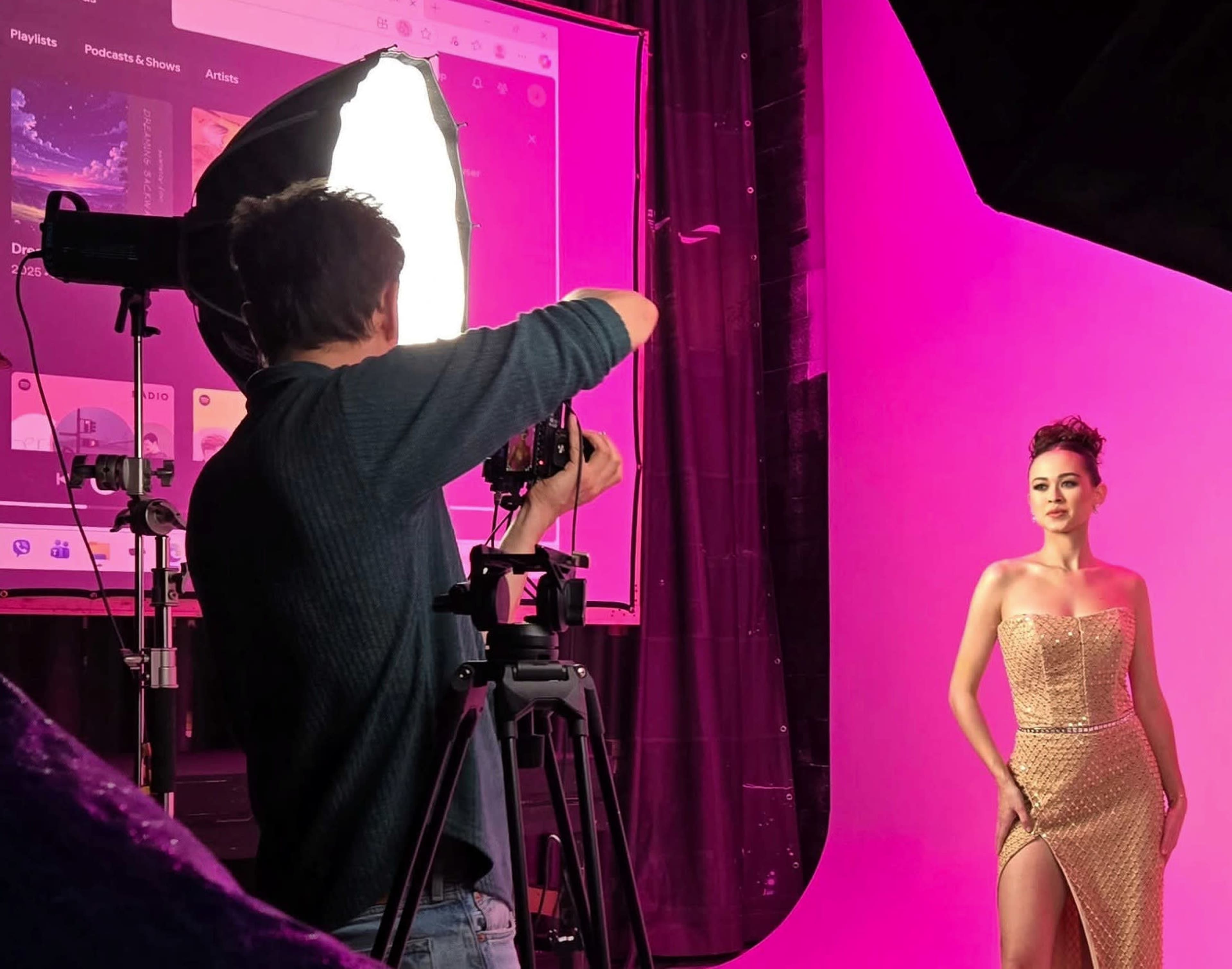 A photographer captures a model posing in a shimmering gold dress against a vibrant pink backdrop in a studio setting.