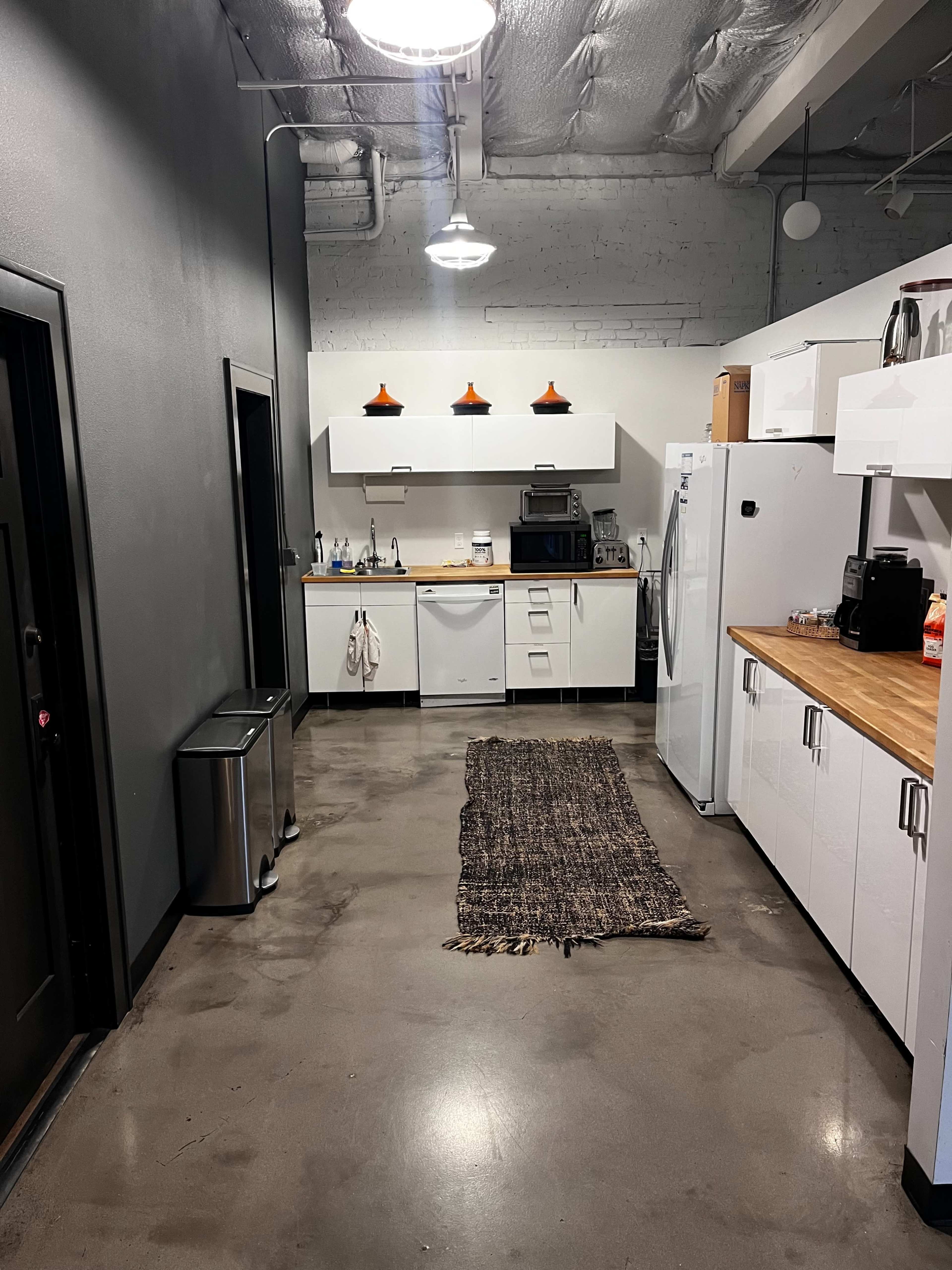 The image shows a modern kitchen area with a long rug running along the polished concrete floor, featuring white cabinets, stainless steel appliances, and pendant lights.