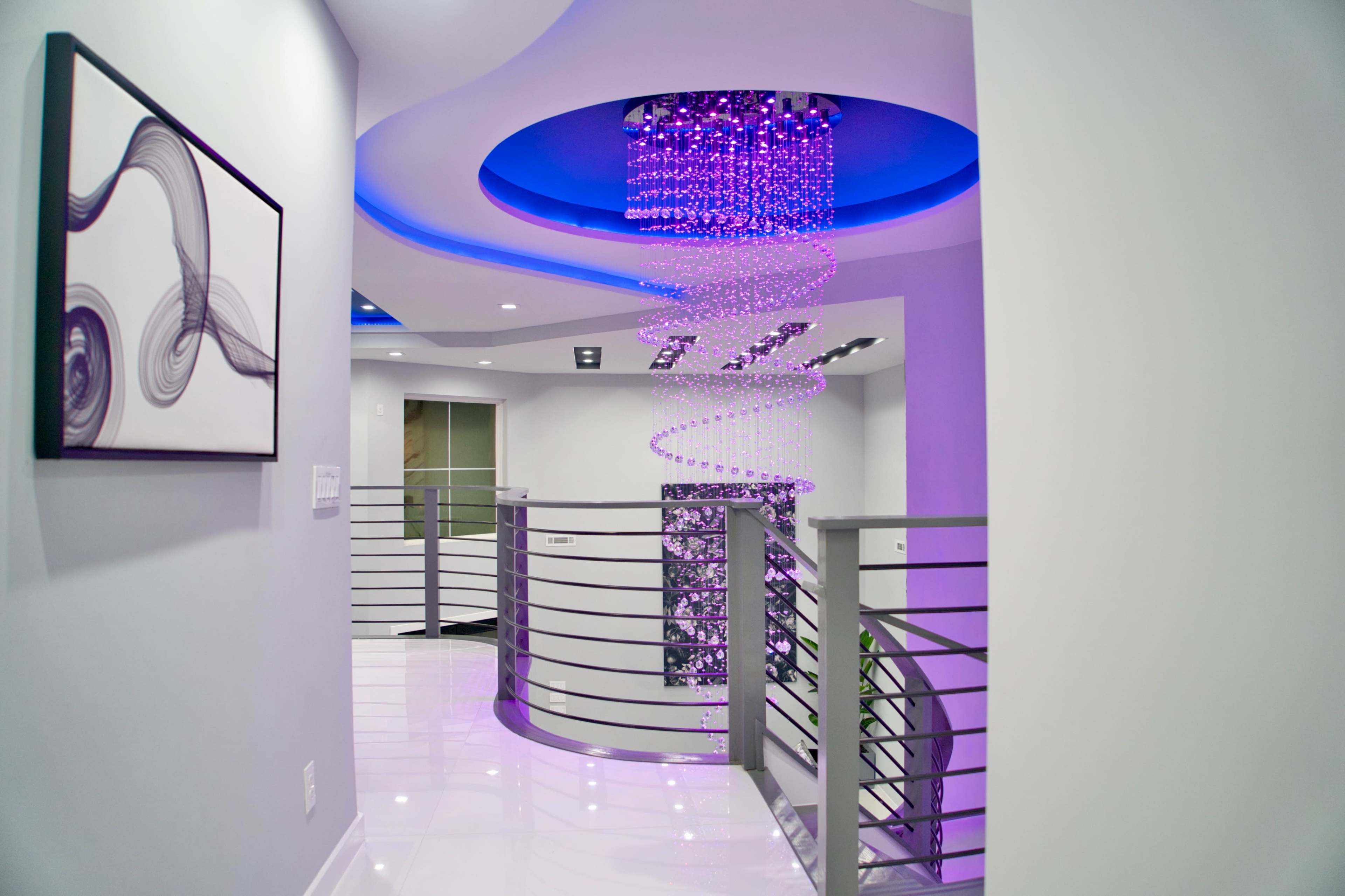A modern hallway features a spiral chandelier with purple lights and a curved railing, complemented by a framed abstract artwork on the wall.