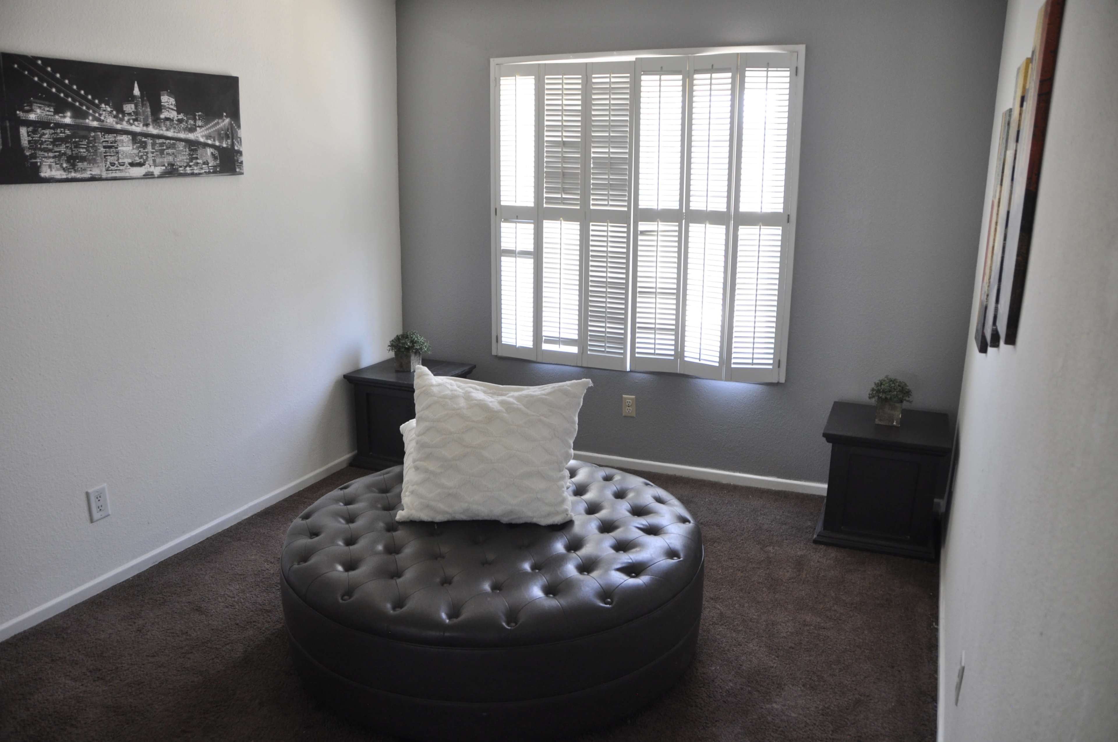 A small room features a tufted circular ottoman in the center, flanked by two dark wooden tables and a window with slatted blinds.