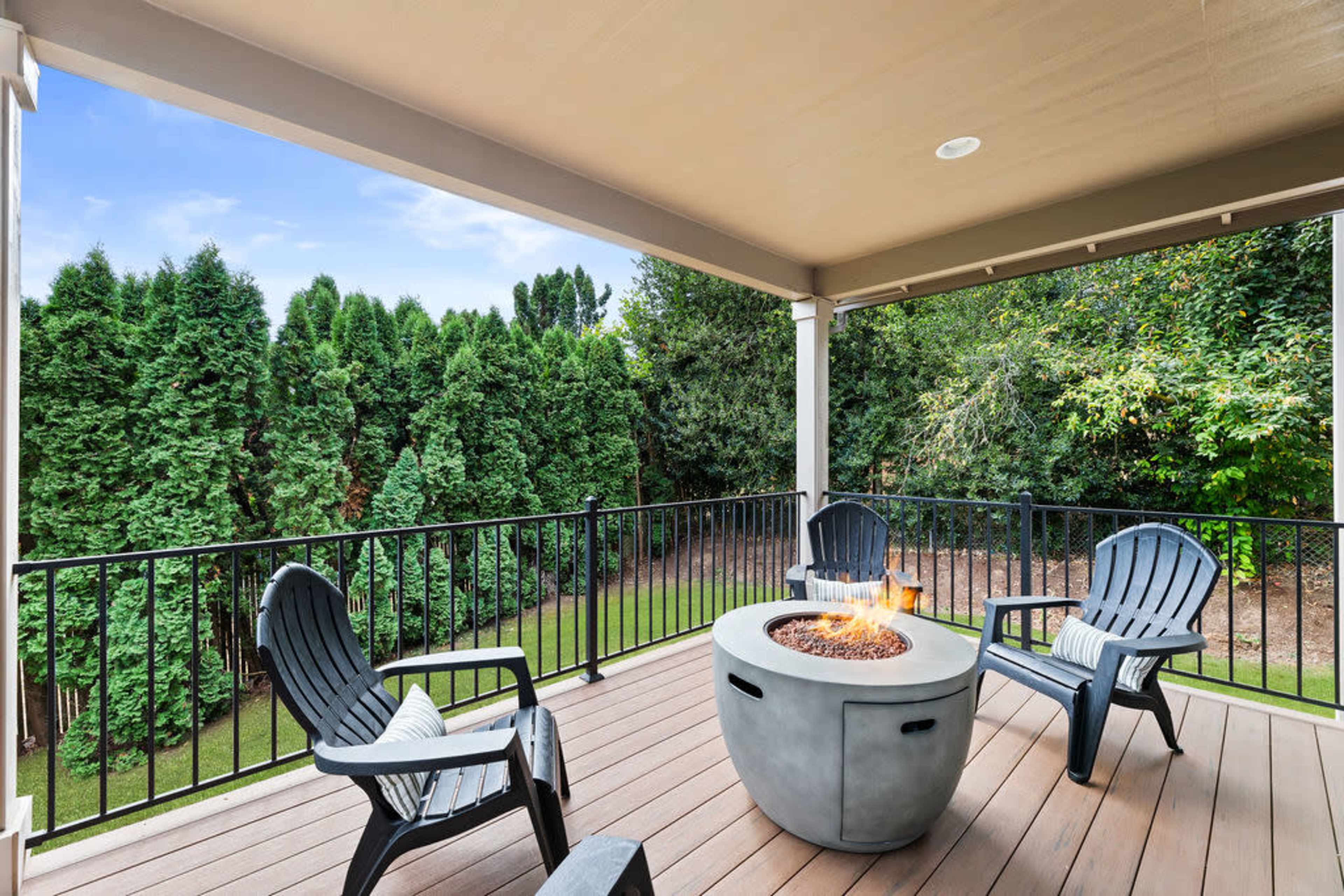 A covered balcony features three black Adirondack chairs arranged around a fire pit, with a green backyard visible in the background.