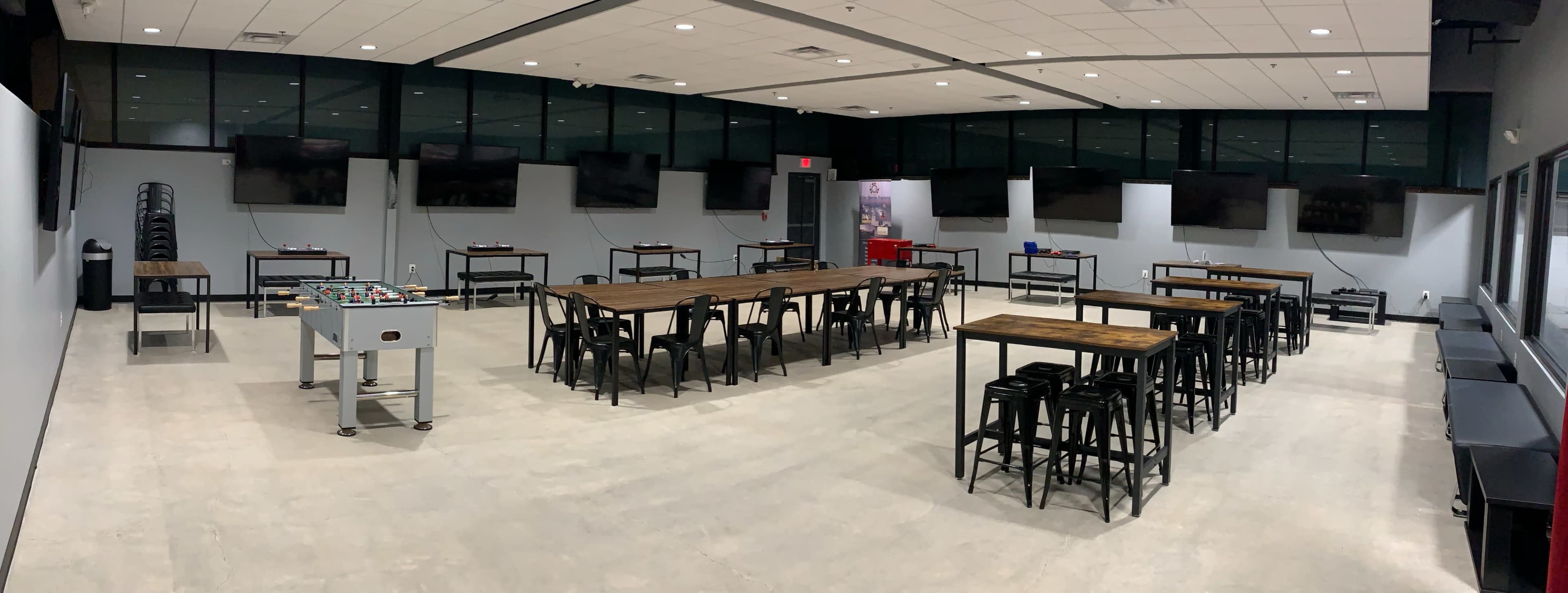 The image shows a spacious room with several long tables and chairs, along with a foosball table and large screens mounted on the walls.