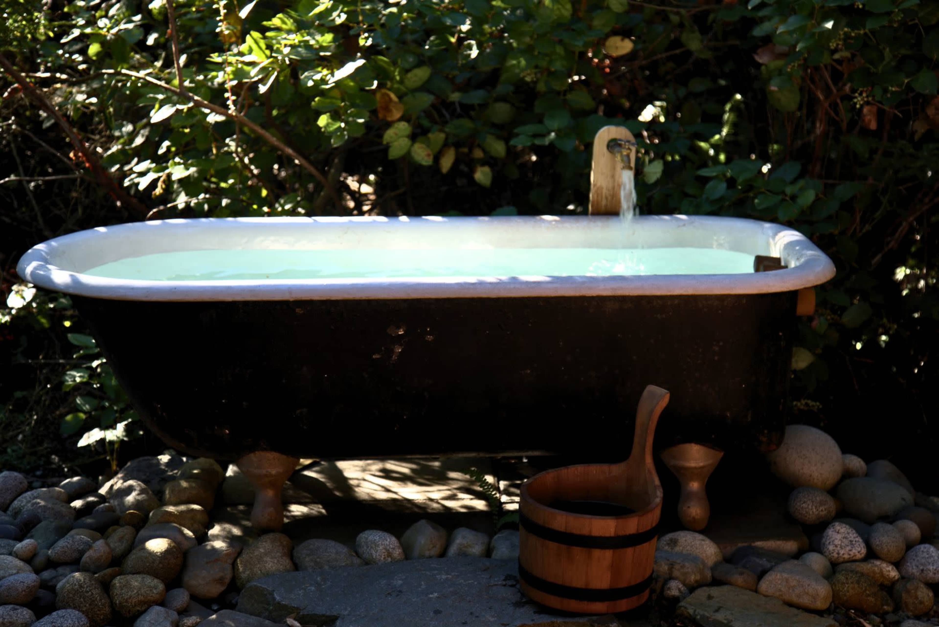 A dark metal bathtub sits on a stone path surrounded by greenery, with water flowing from a spout into the tub and a wooden bucket beside it.
