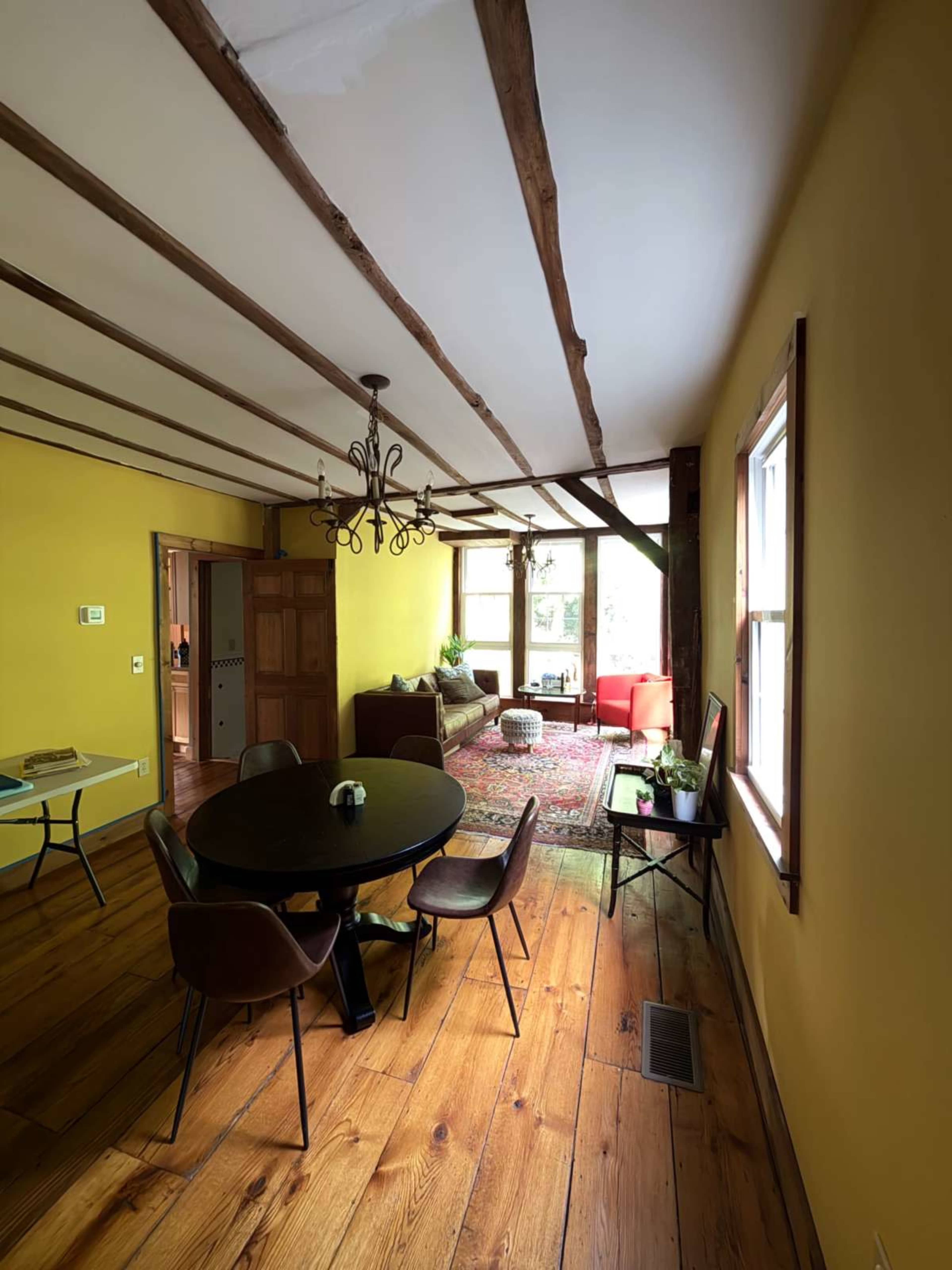 The image shows a cozy dining area with a round table and chairs, leading into a room with yellow walls, wooden beams, and a small seating area.