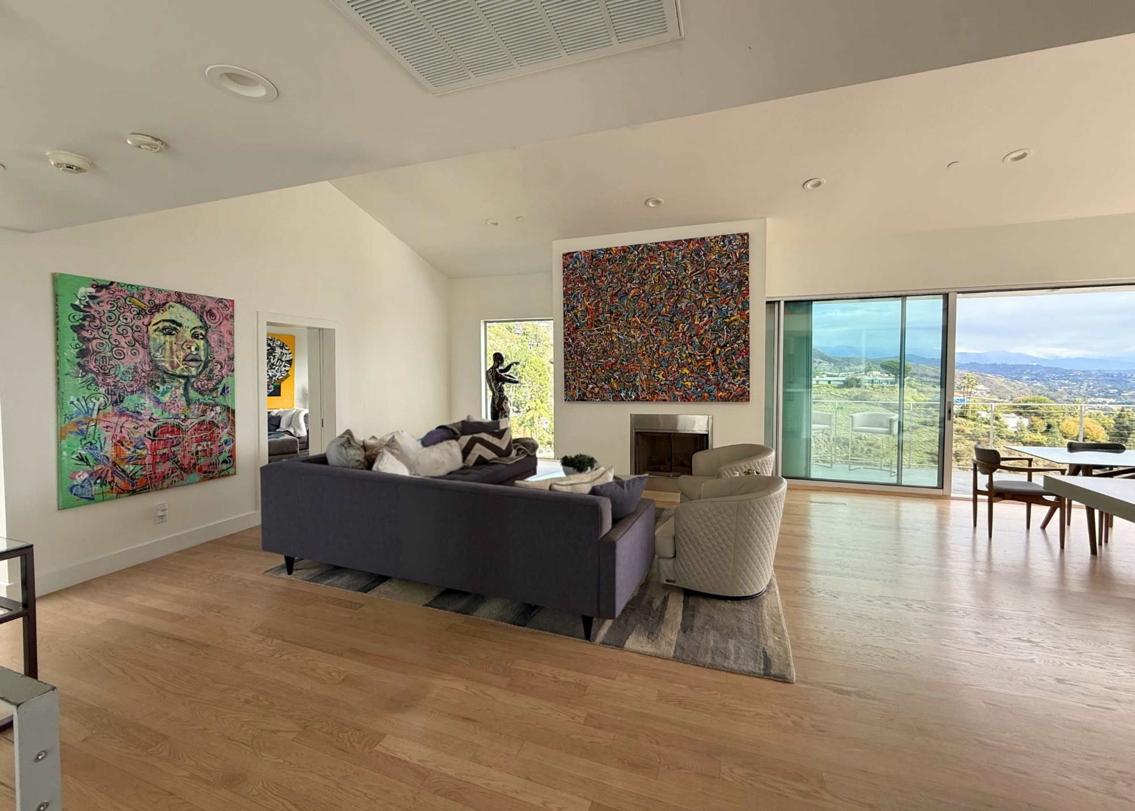 The interior of a modern living room features a dark sectional sofa, two light armchairs, and colorful artwork on the walls, with large windows showcasing a mountain view.
