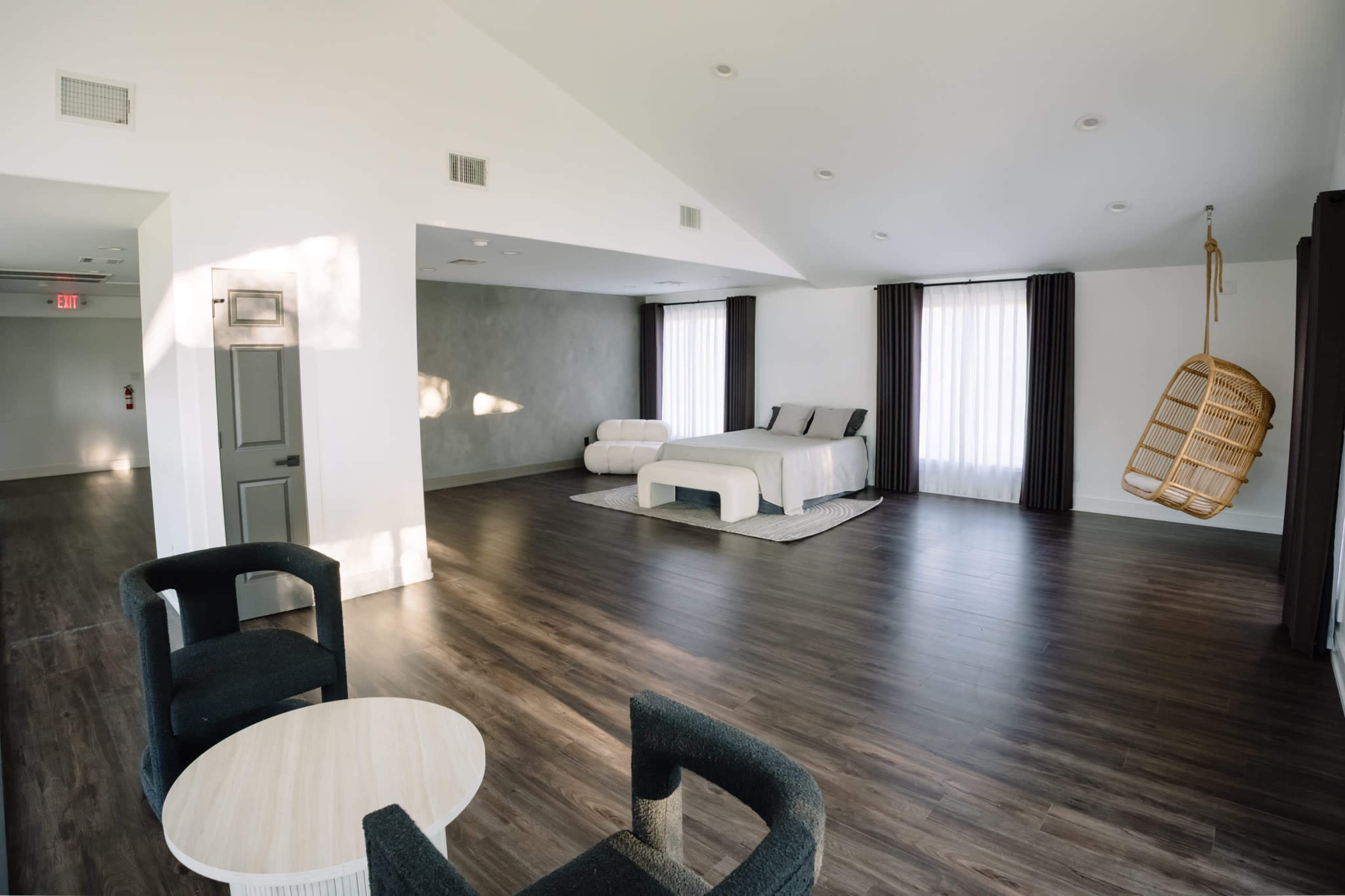 The image shows a spacious, modern bedroom featuring a large bed, a hanging chair, and minimalistic furniture, all set against a backdrop of wooden flooring and large windows.