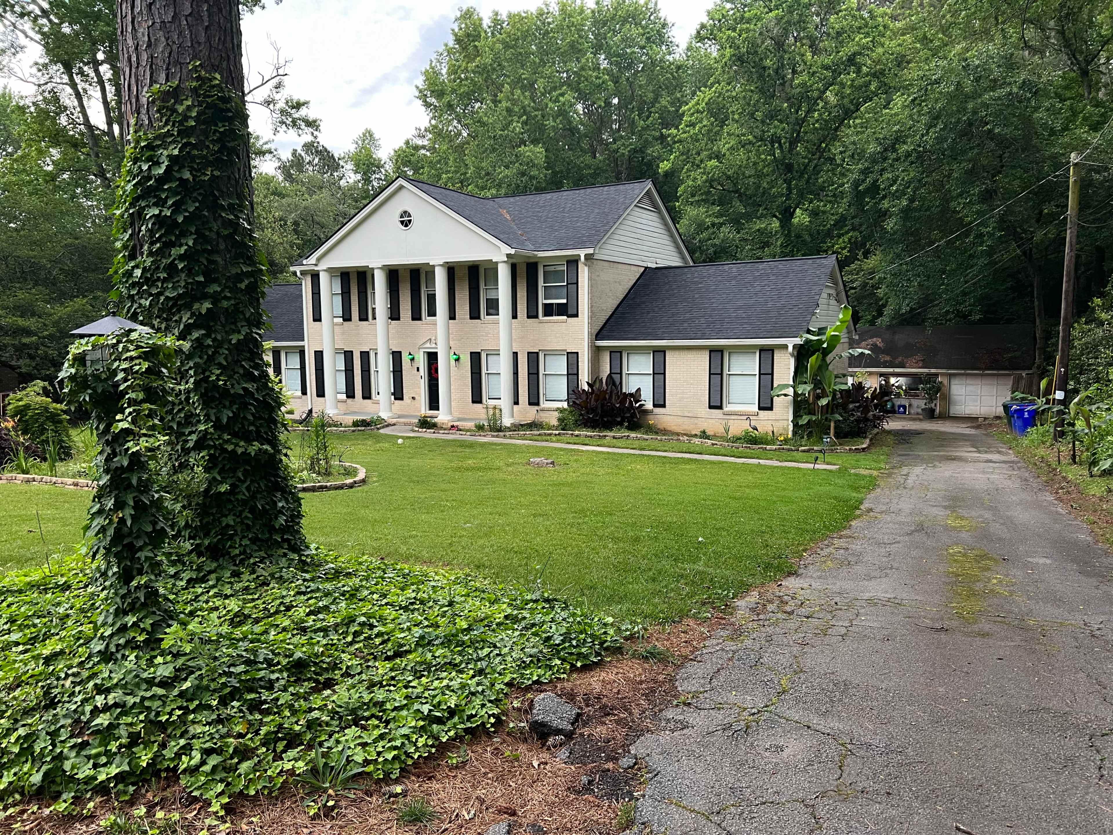 A two-story house with a light-colored facade and a large front lawn is surrounded by trees and a gravel driveway.