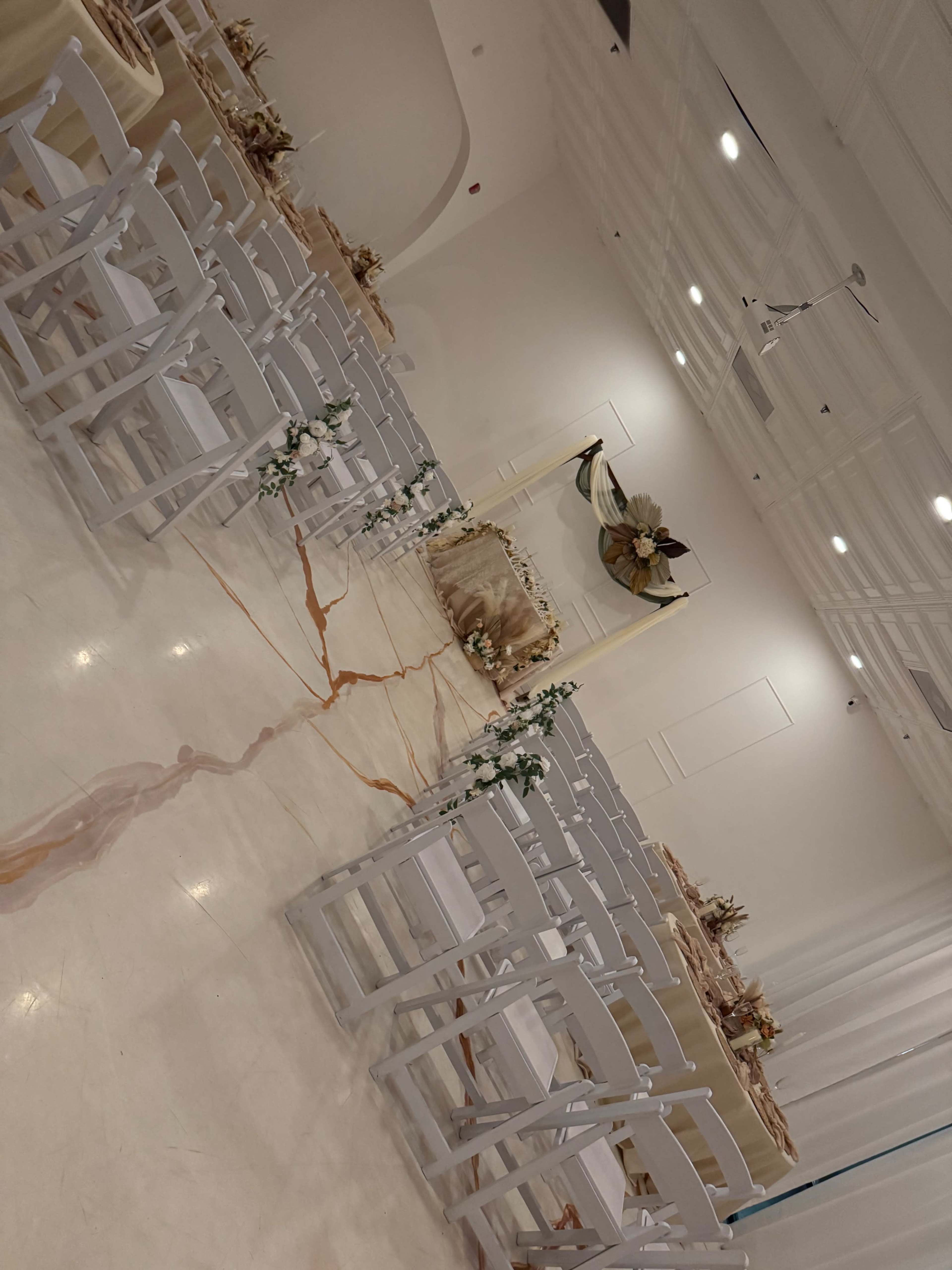 The image shows a decorated indoor venue set up for an event, with rows of white chairs facing a stage adorned with floral arrangements and draped fabric.