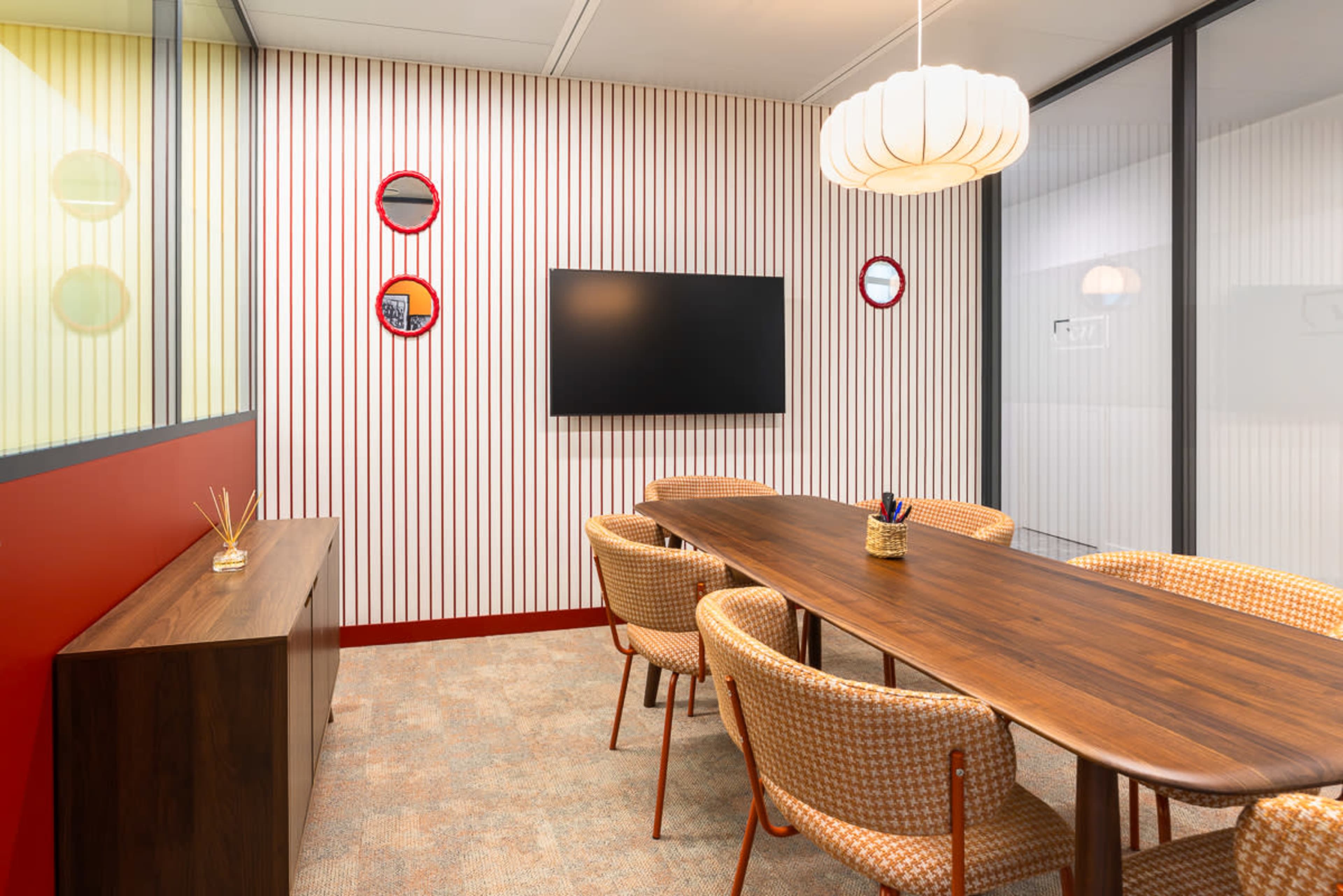 A modern conference room features a long wooden table surrounded by wicker chairs, with a large screen mounted on a striped wall and decorative mirrors.