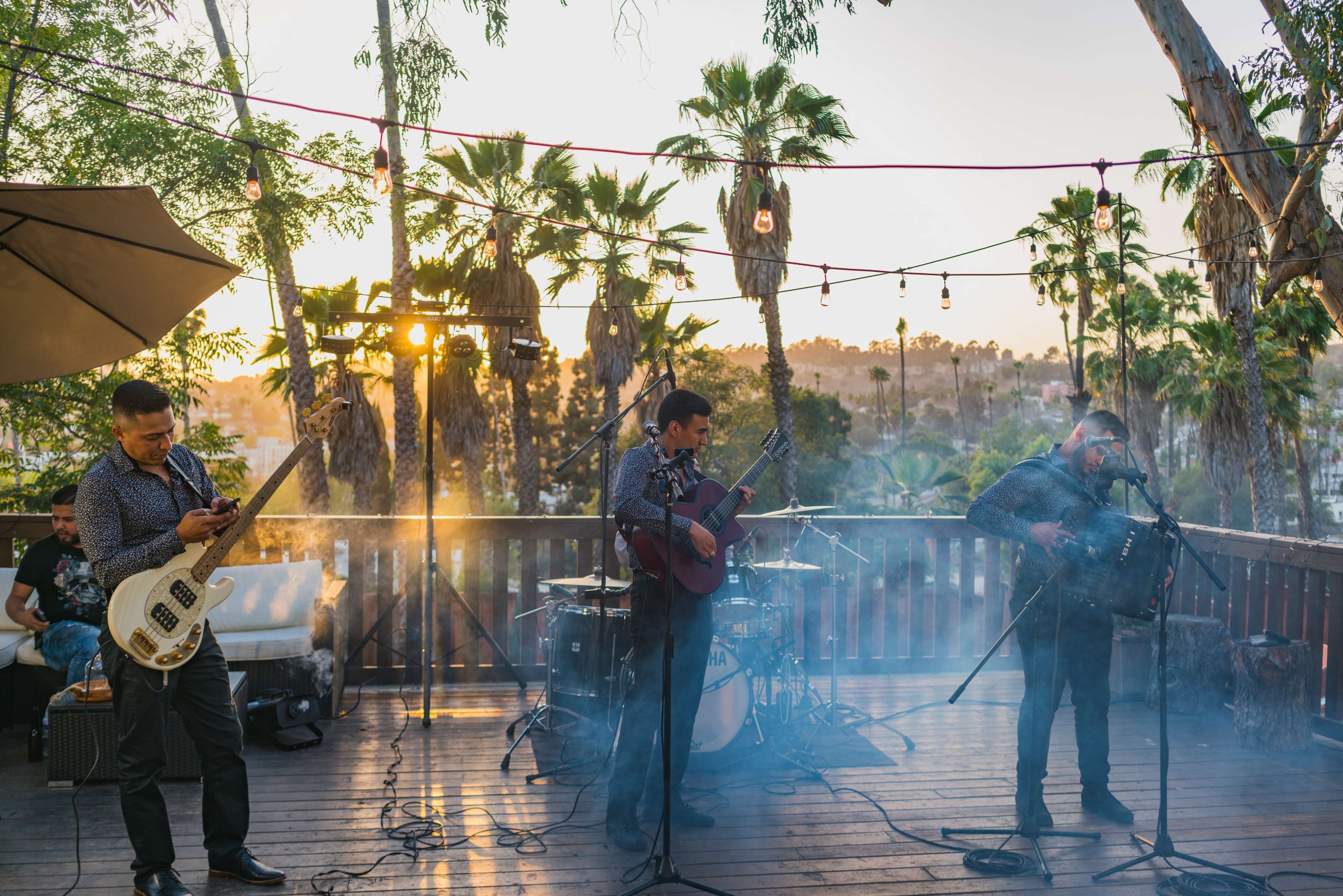 Three musicians perform on a wooden deck surrounded by palm trees at sunset, with lights strung overhead.