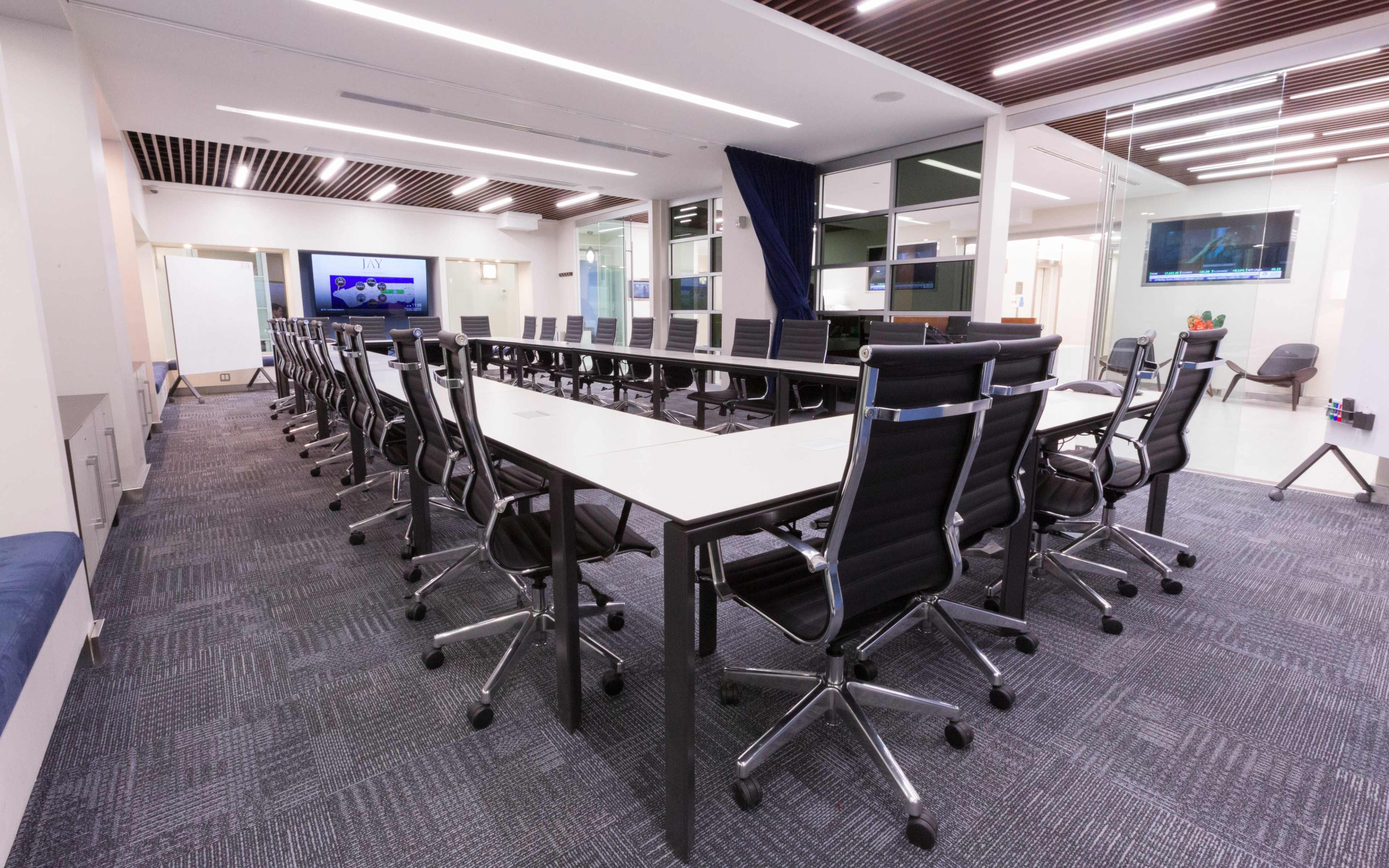 A large, modern conference room with a long table surrounded by black ergonomic chairs and glass walls, featuring electronic displays.