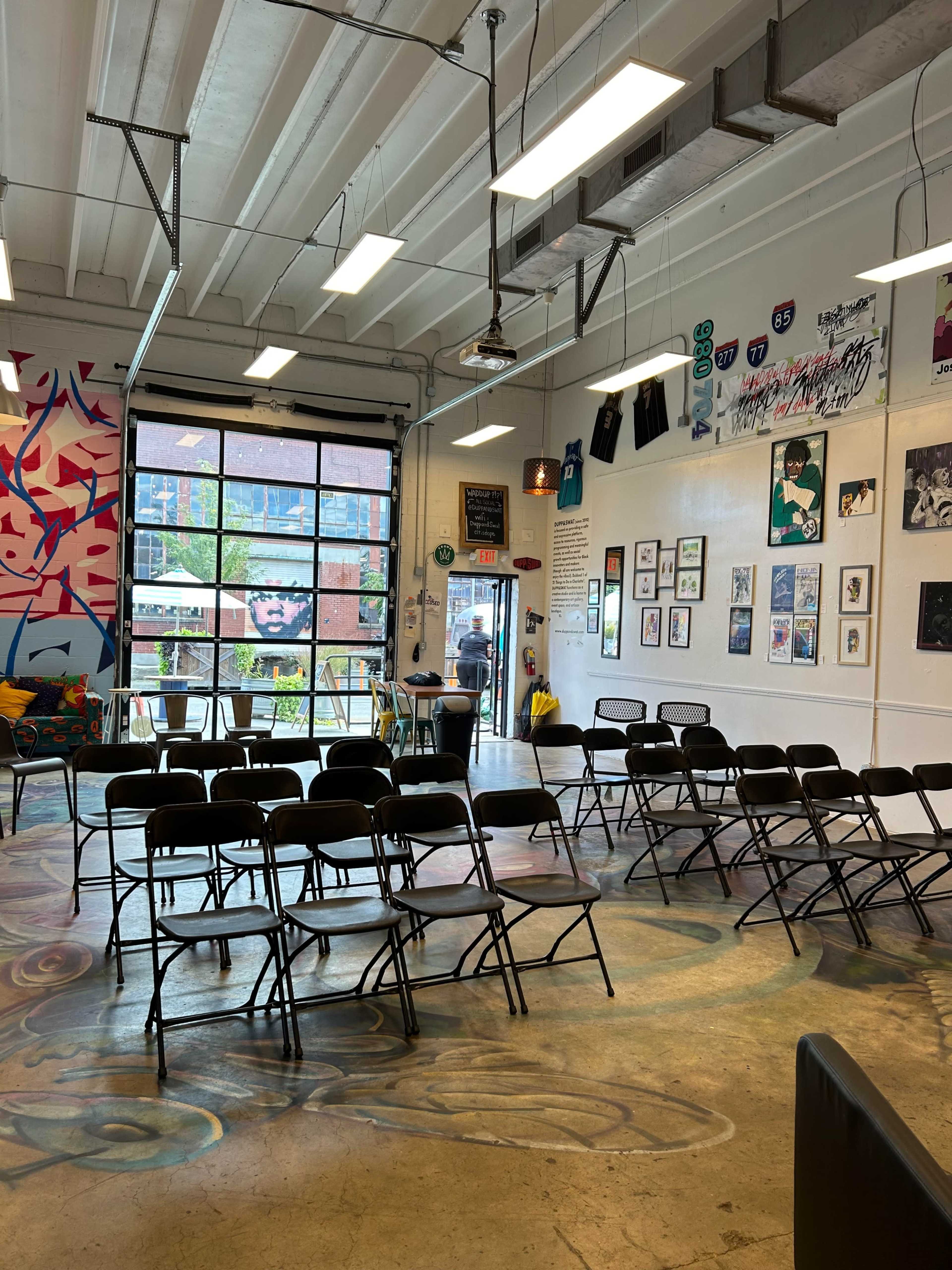 The image shows a brightly lit room with folding chairs arranged in rows, colorful wall art, and large windows providing natural light.