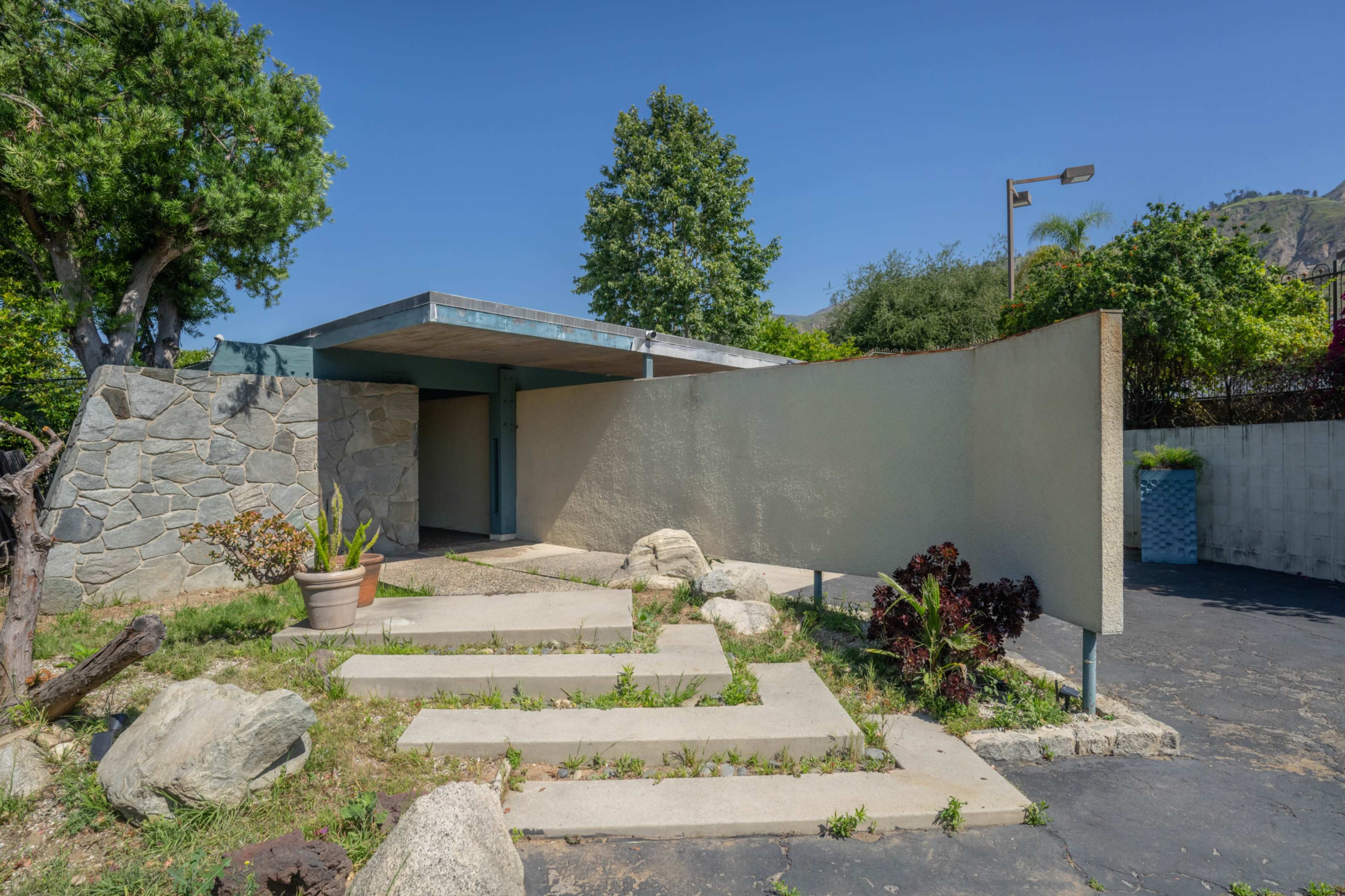 The image shows a modern house with a flat roof and a stone pathway leading to the entrance, surrounded by greenery and rocks.