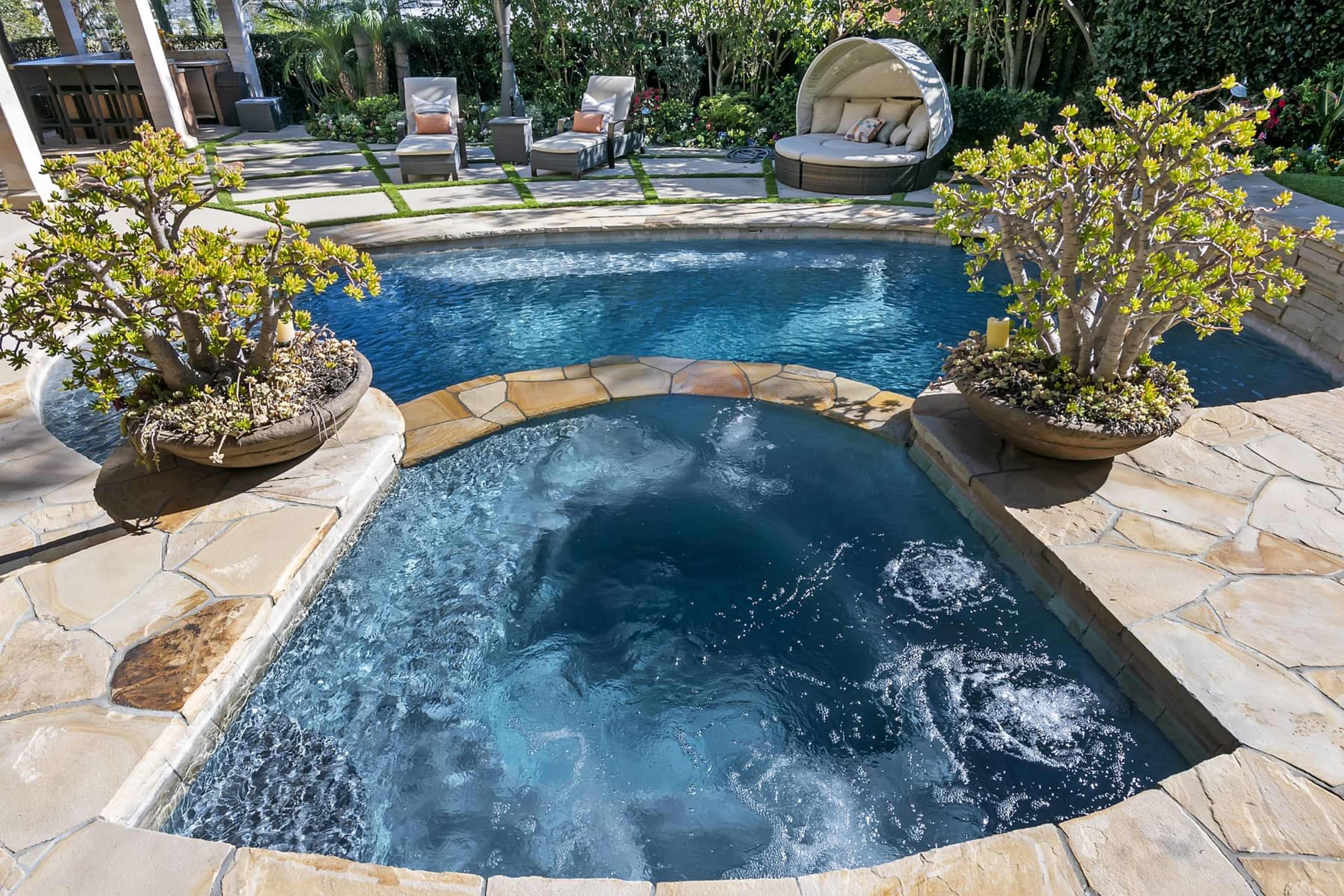 A circular hot tub is situated next to a rectangular swimming pool, surrounded by leafy plants and lounge chairs.