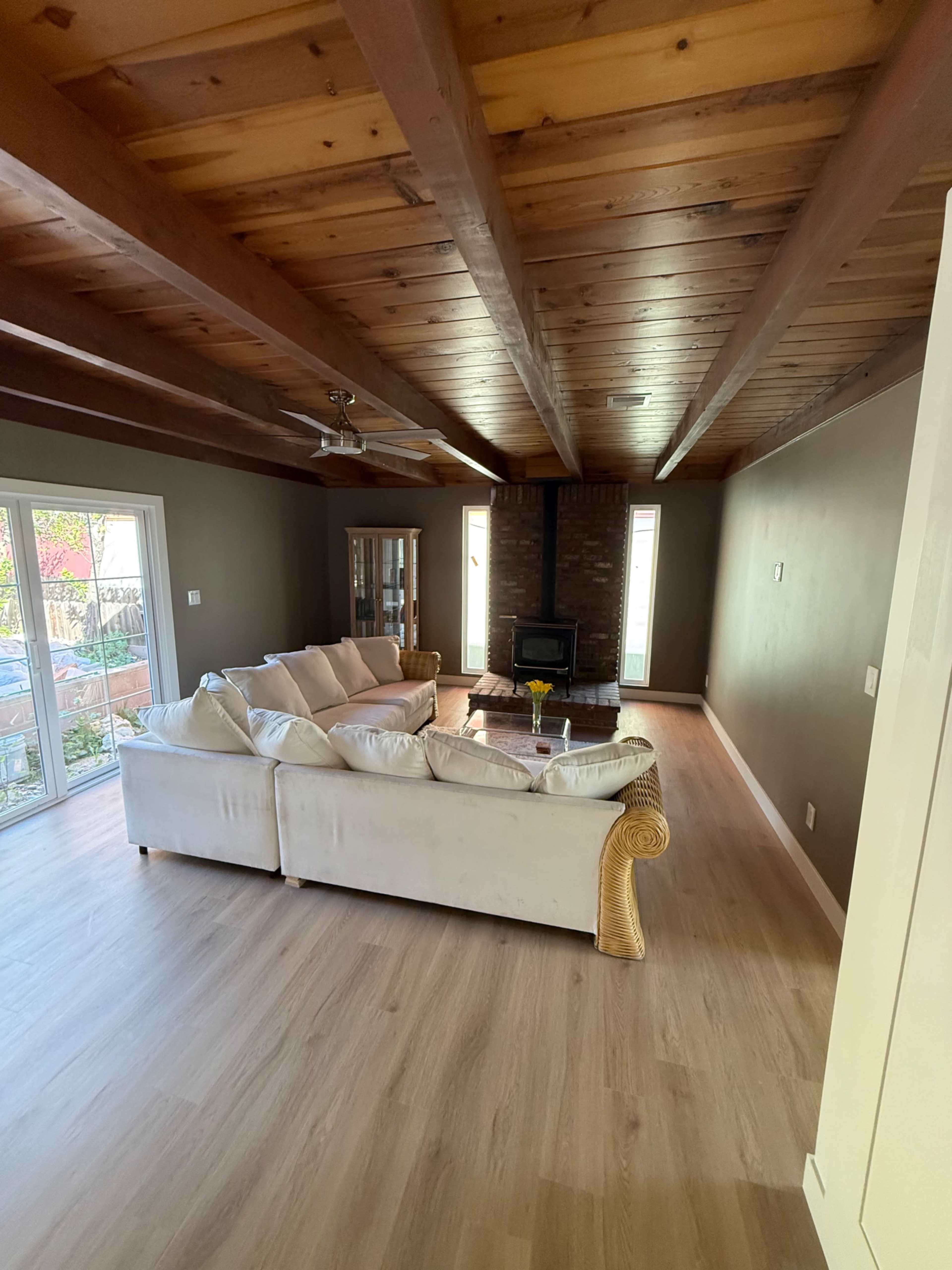 The image shows a spacious living room with a beige sectional sofa, wooden beams on the ceiling, and a stone fireplace, featuring large windows that provide natural light.